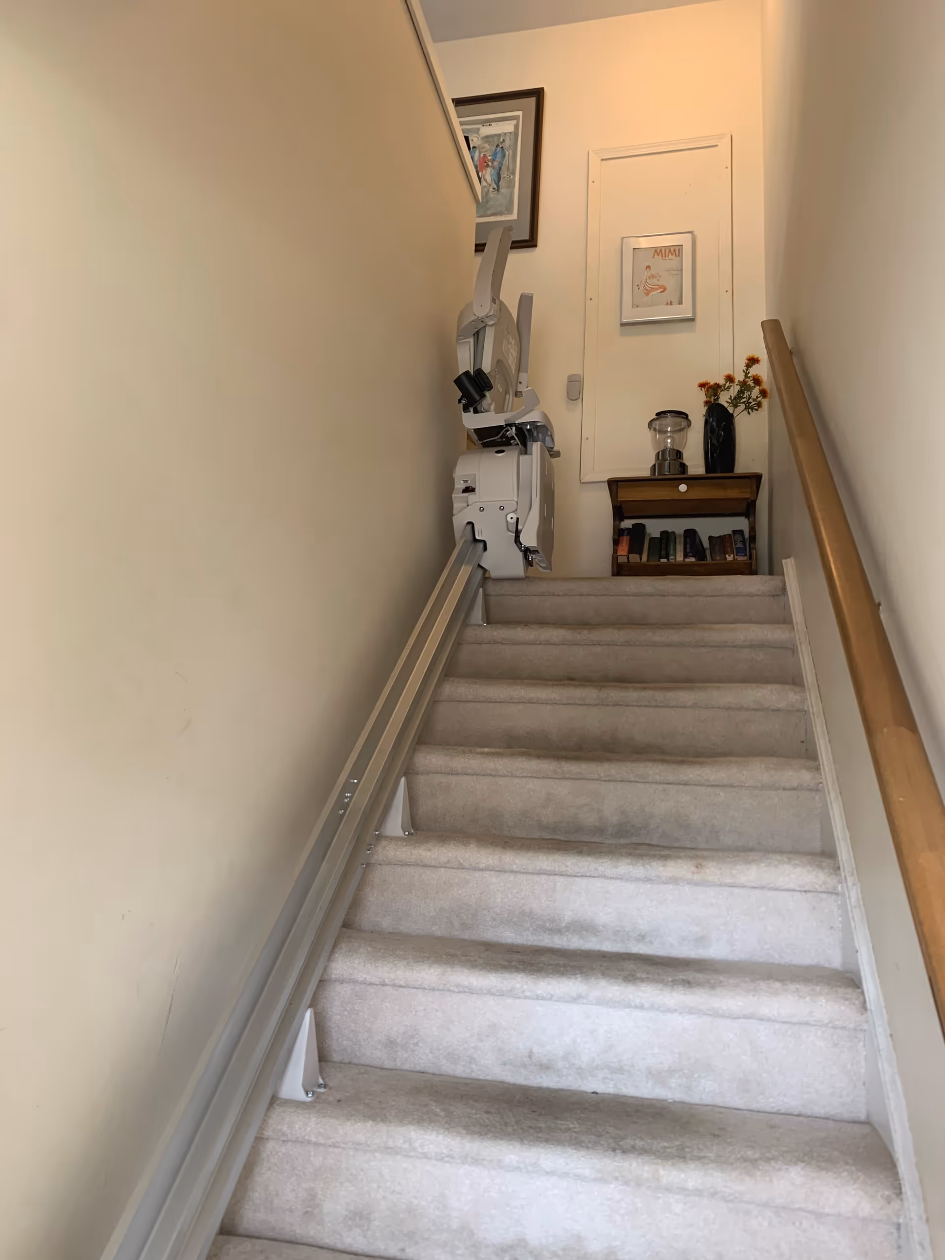 Narrow carpeted stairwell with a mounted chairlift and small cabinet at the top.