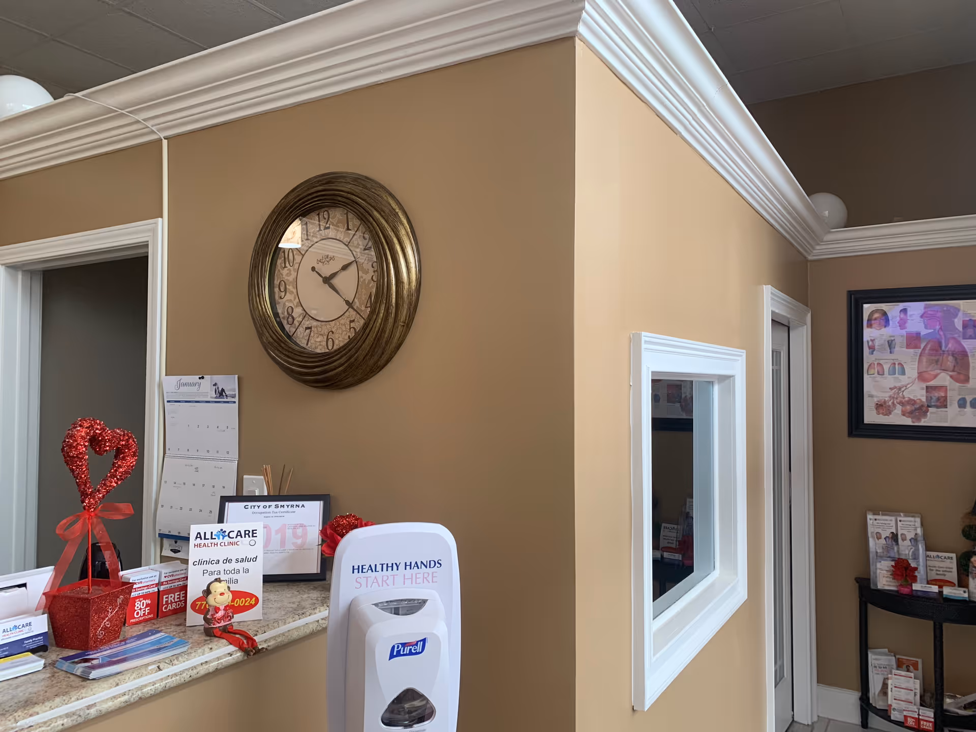 Reception area with beige walls, a large wall clock, brochures, and a hand sanitizer dispenser.