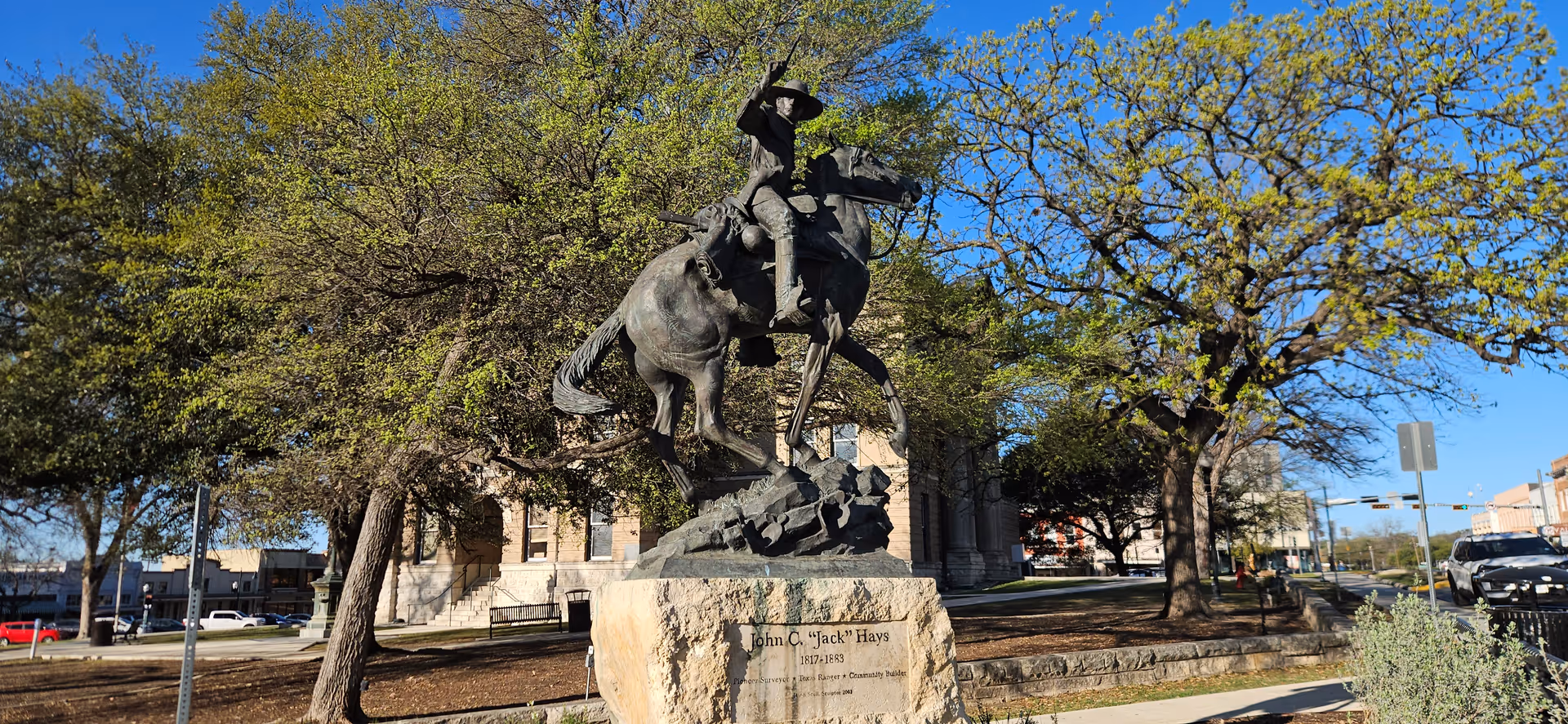 Bronze statue of a rider on a rearing horse in a park surrounded by trees.