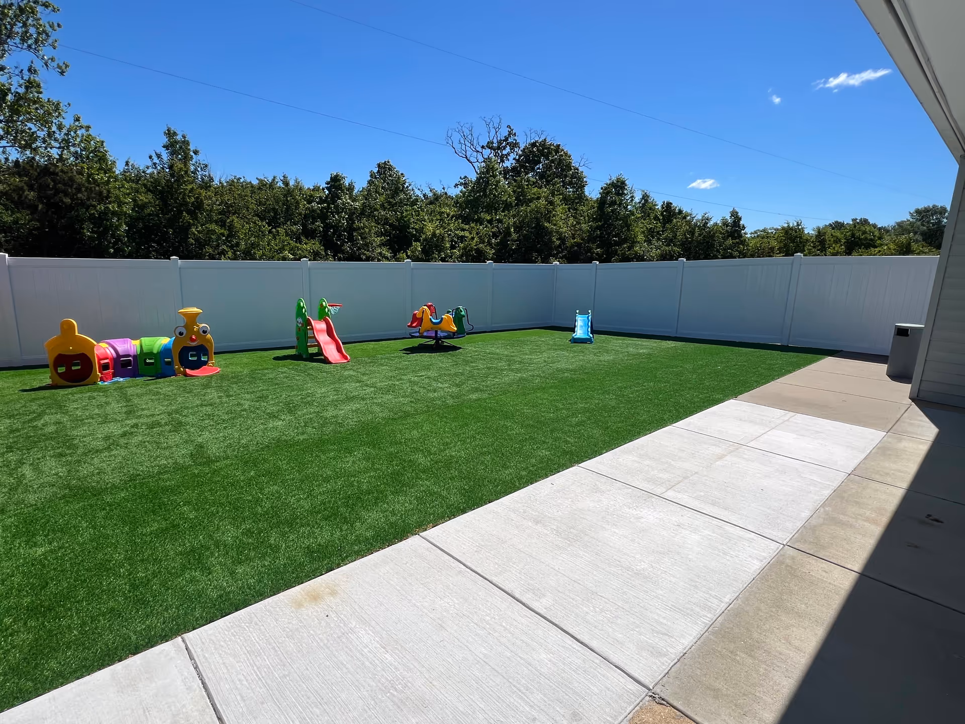 Fenced outdoor play area with colorful plastic toys and a green artificial lawn.