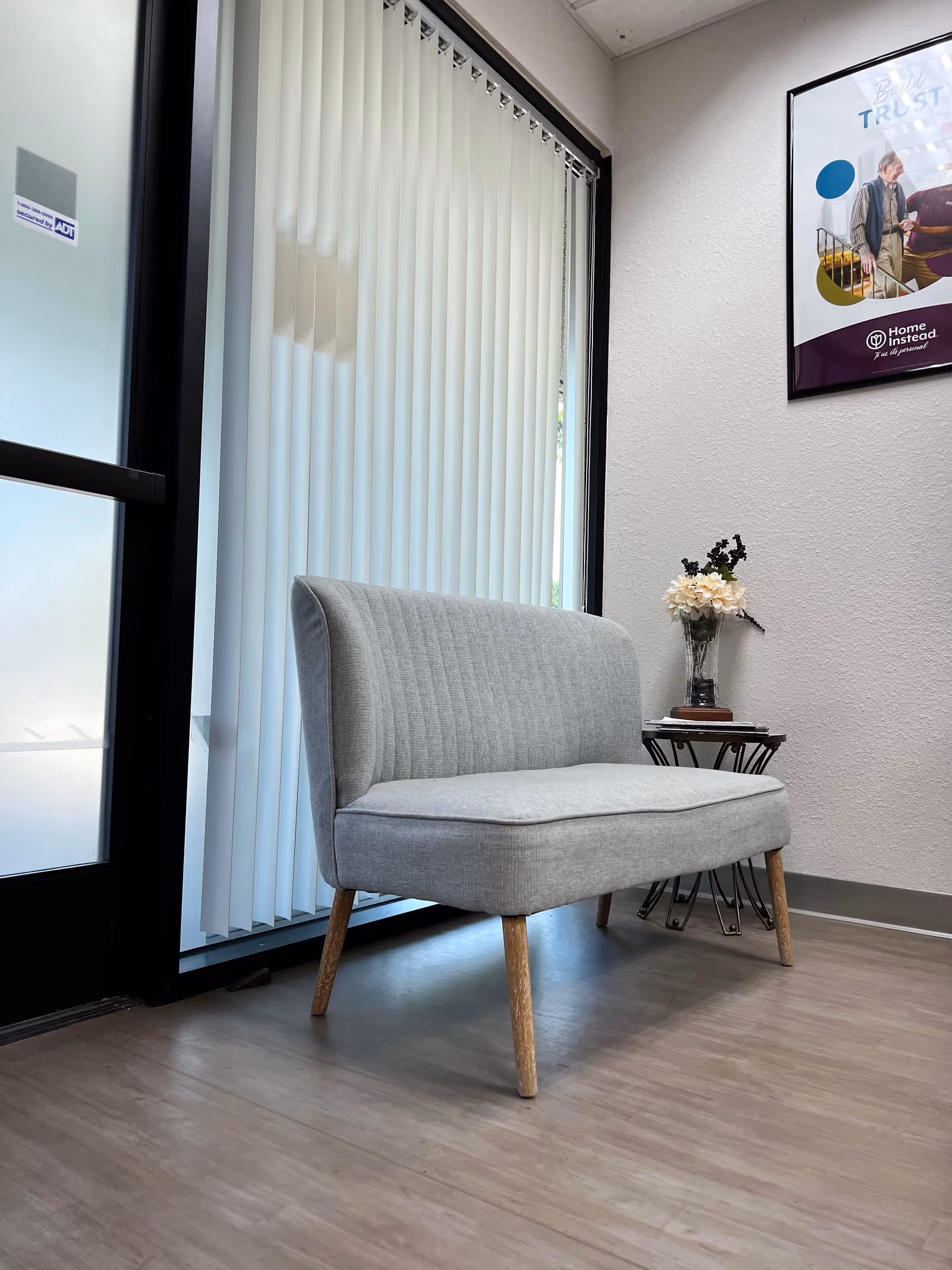 Grey upholstered chair beside vertical blinds, small table with flowers in a waiting lobby.