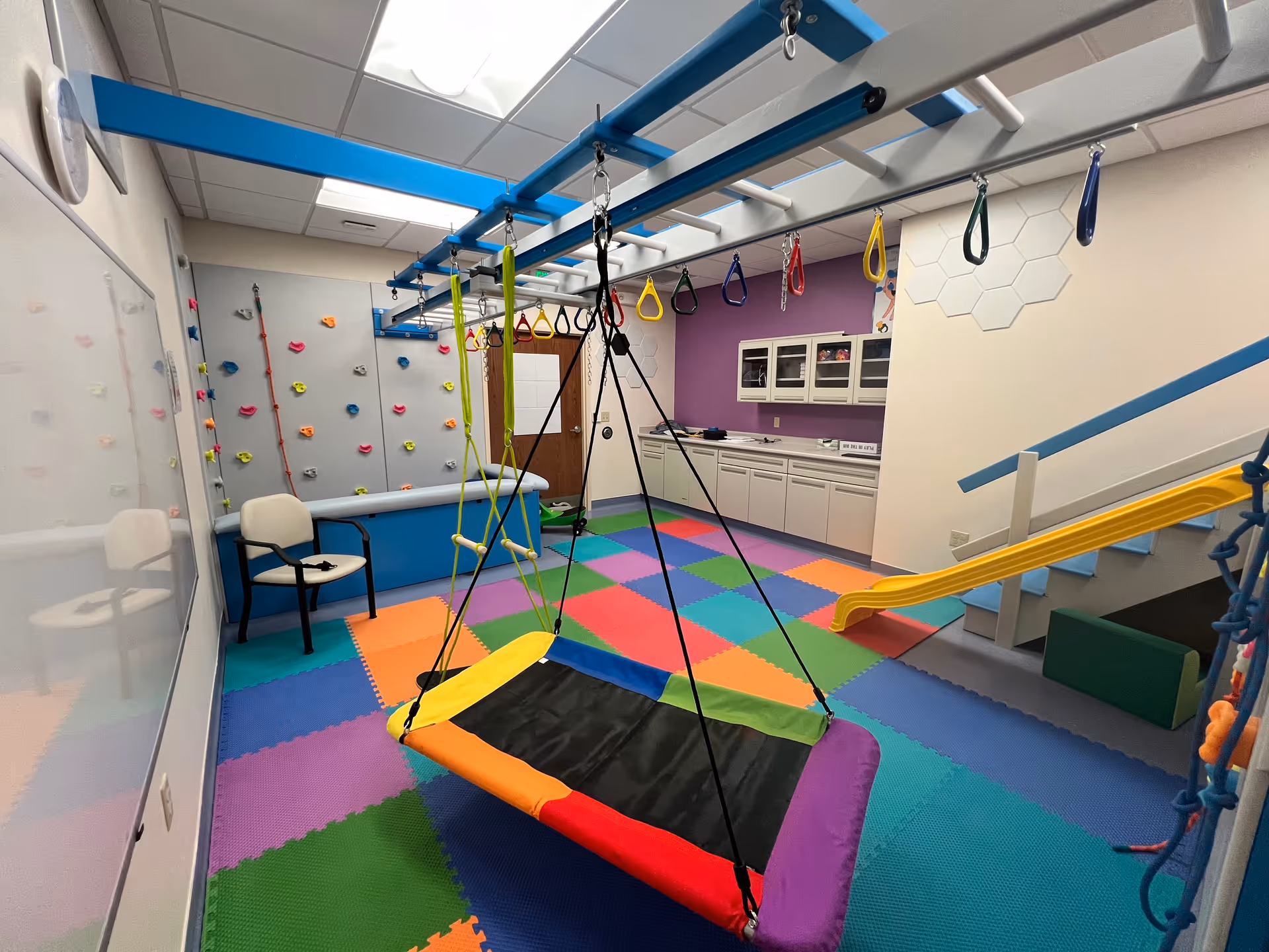 Colorful indoor pediatric therapy playroom with climbing wall, swings, slide, and foam mats.