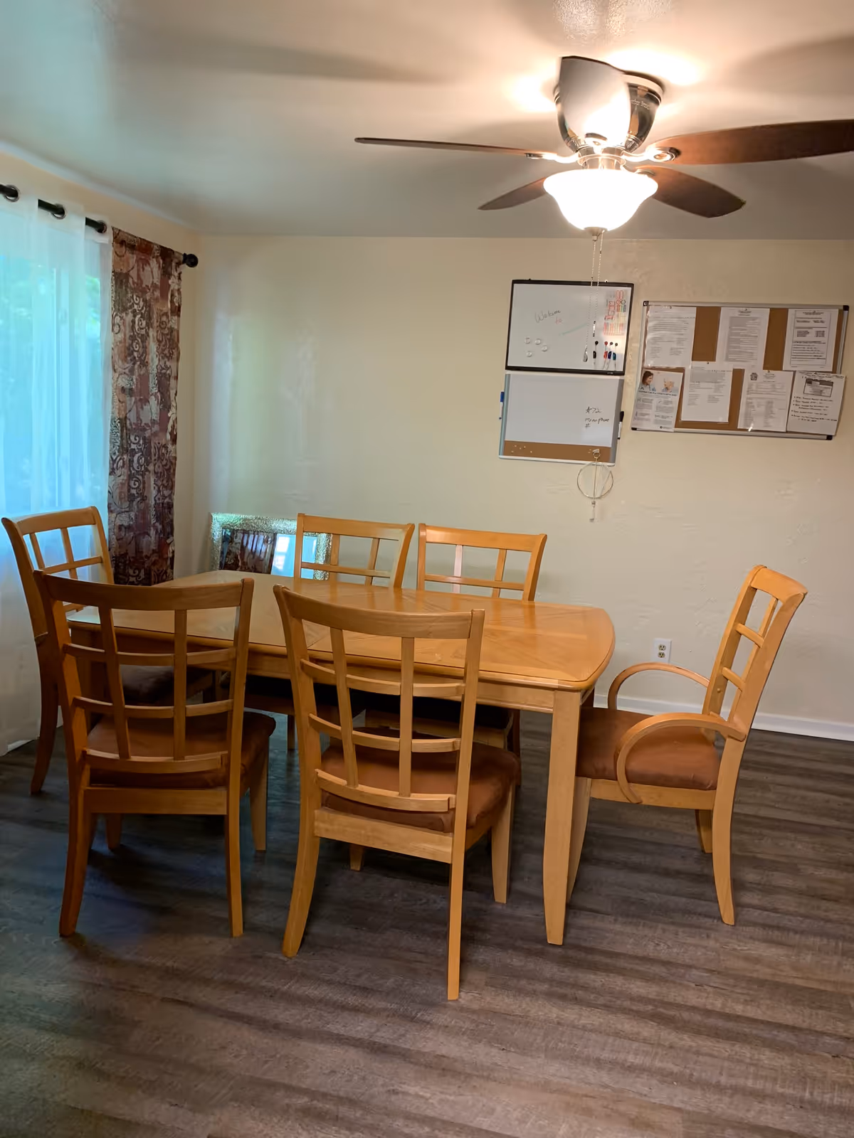 Wooden dining table with chairs in a bright, cozy room with a ceiling fan.