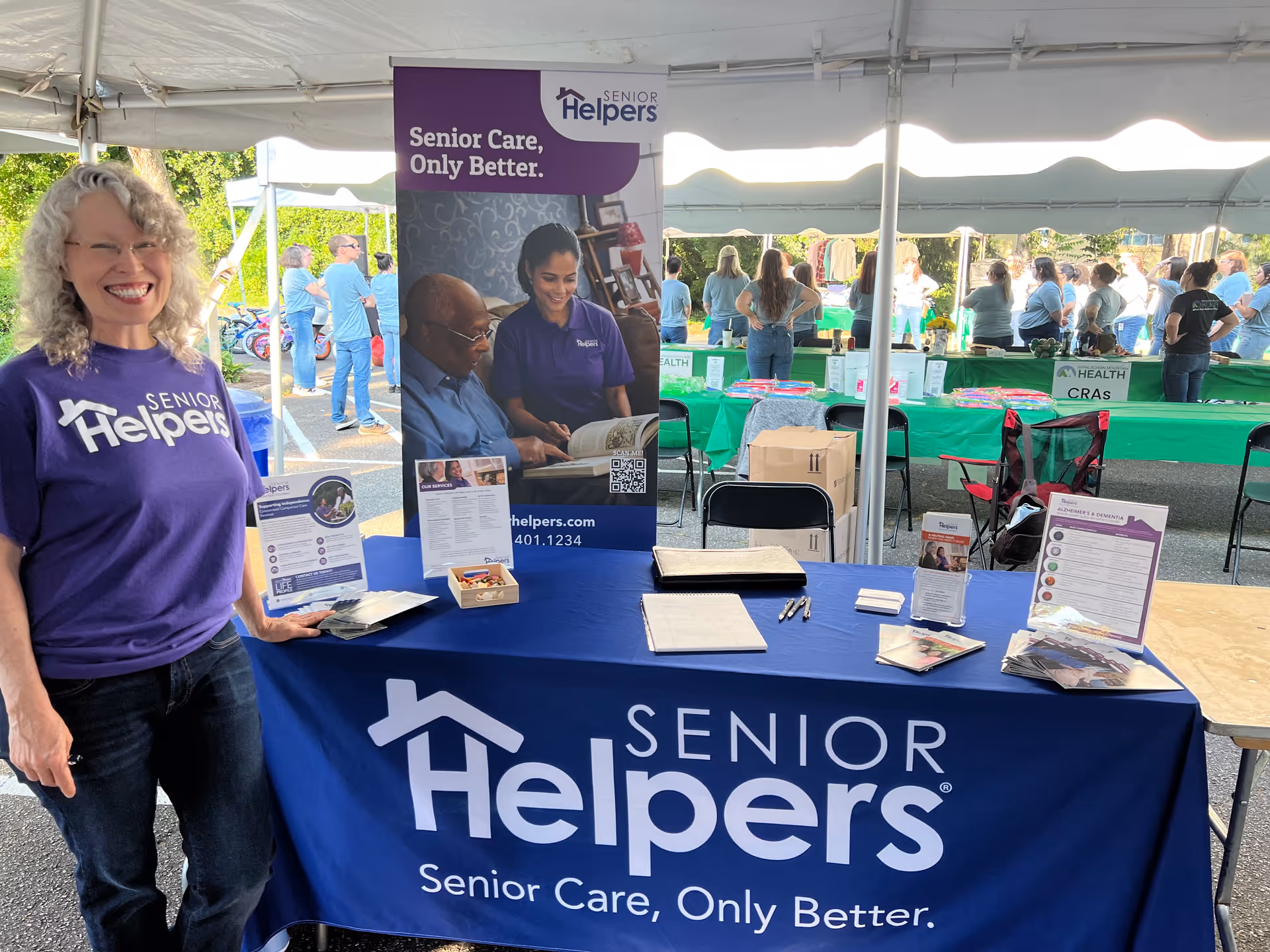Smiling woman in purple Senior Helpers shirt stands beside a booth under a white event tent.