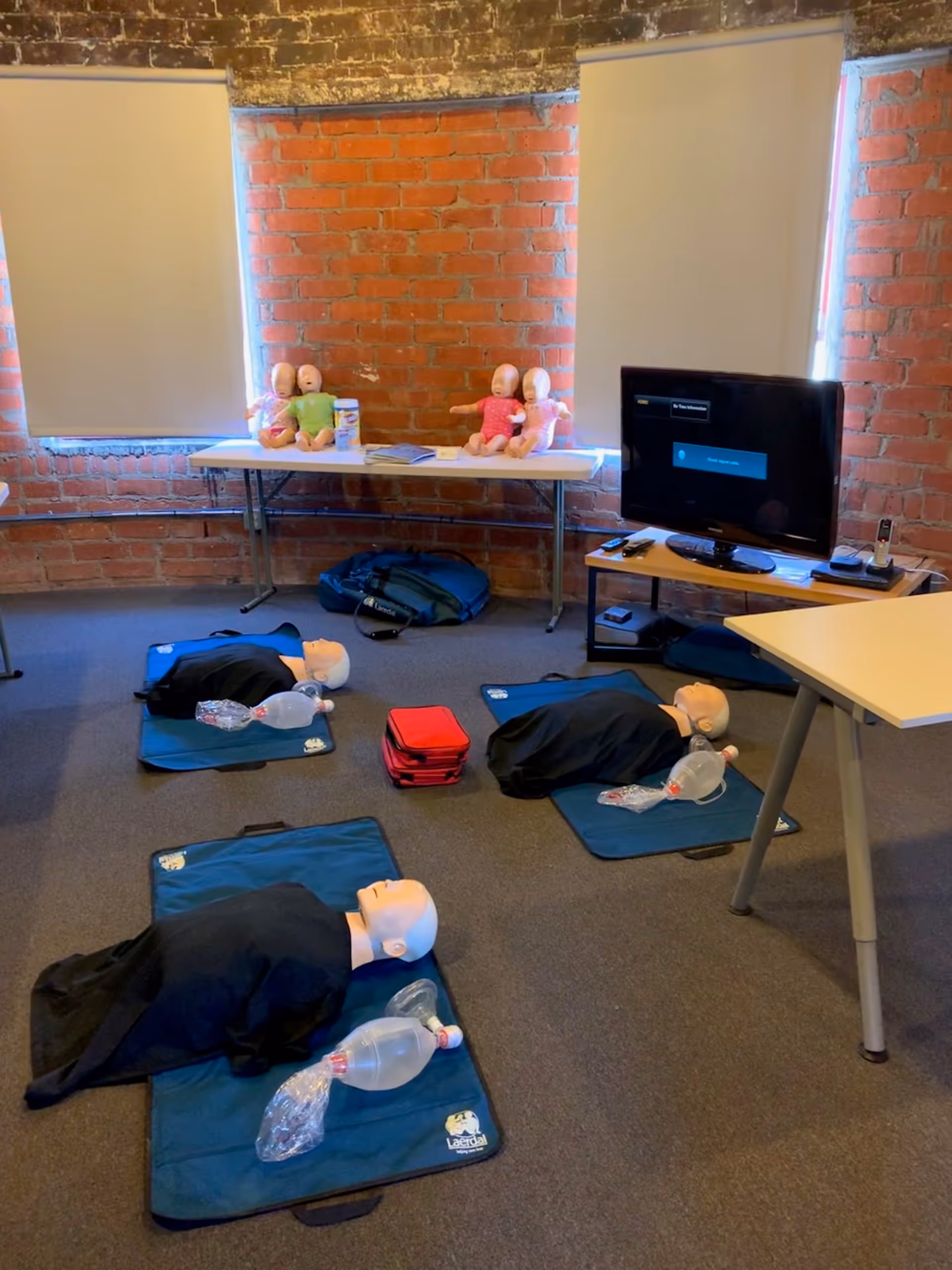 CPR training room with multiple mannequins on mats and a TV against brick walls.