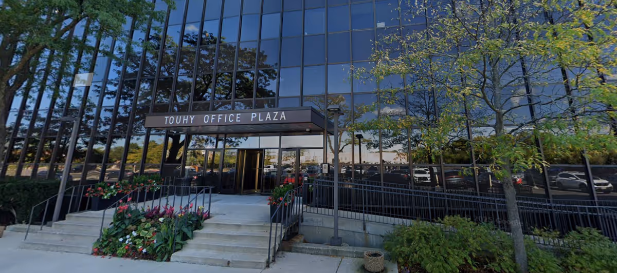 Glass-front office plaza building with stairs, planters, and a shaded entrance.