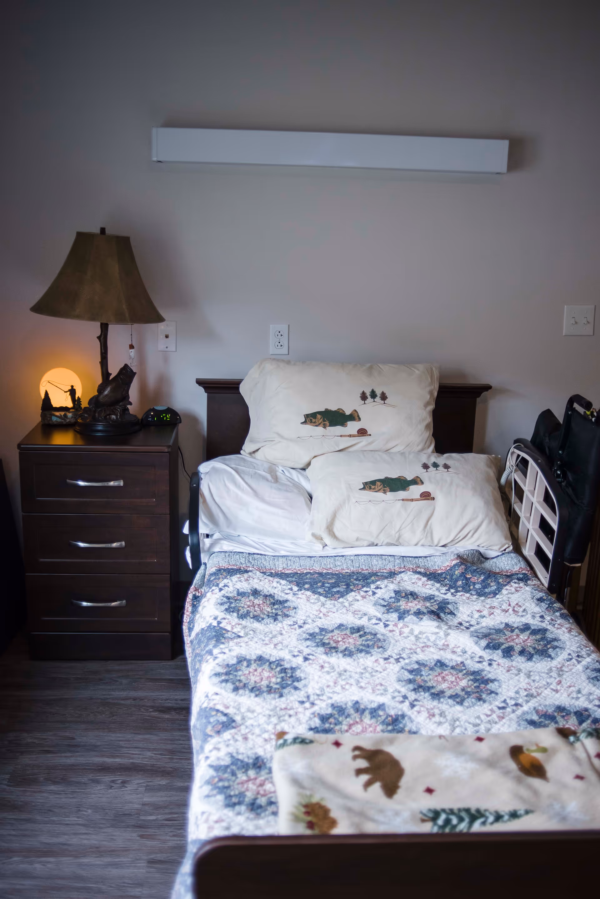A bedroom in a senior facility with a quilted bed, pillows, lamp, and walker.