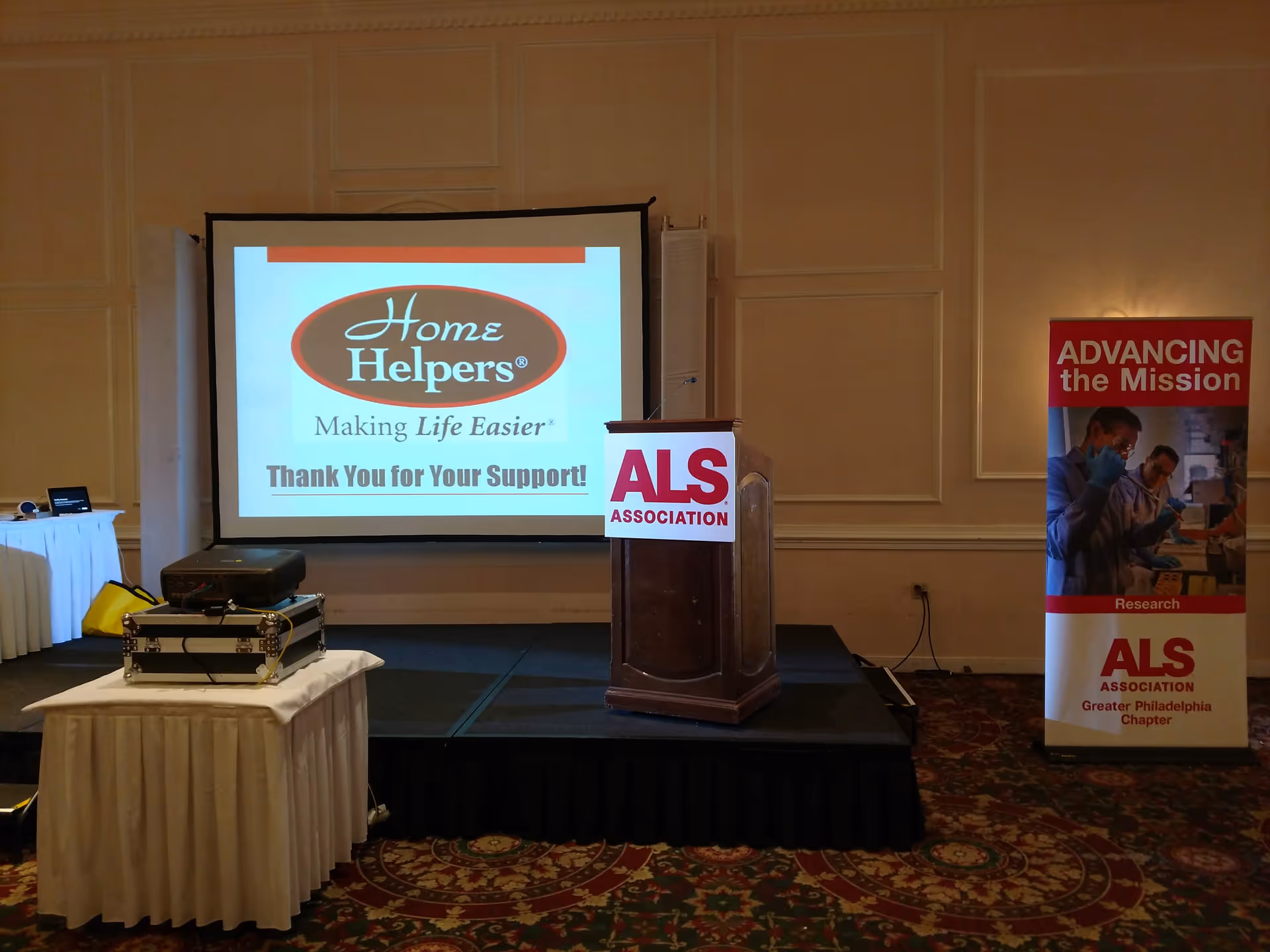 Stage setup with projector screen displaying Home Helpers logo and ALS banners indoors.
