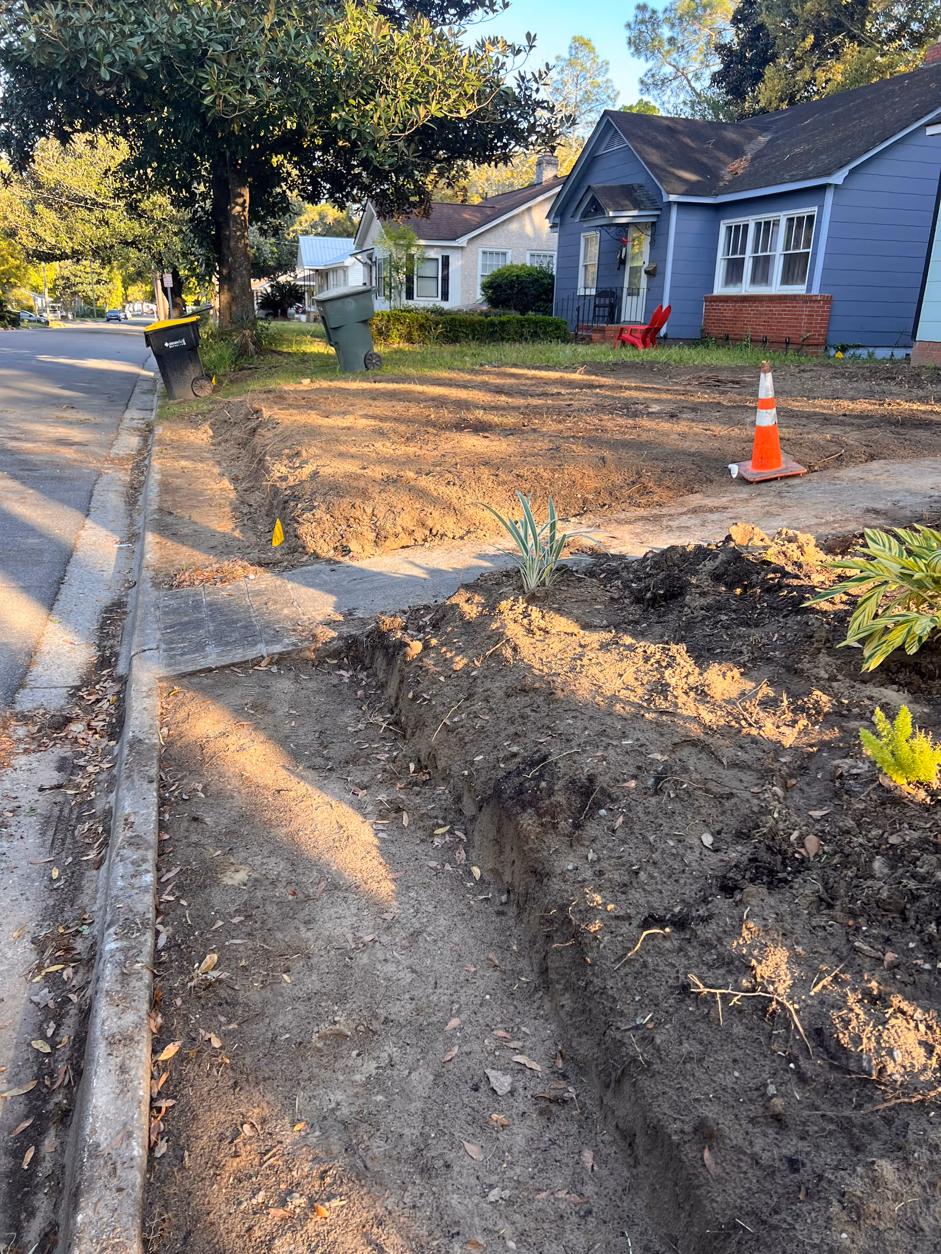 A residential street with a deep trench and an orange traffic cone near a blue house.