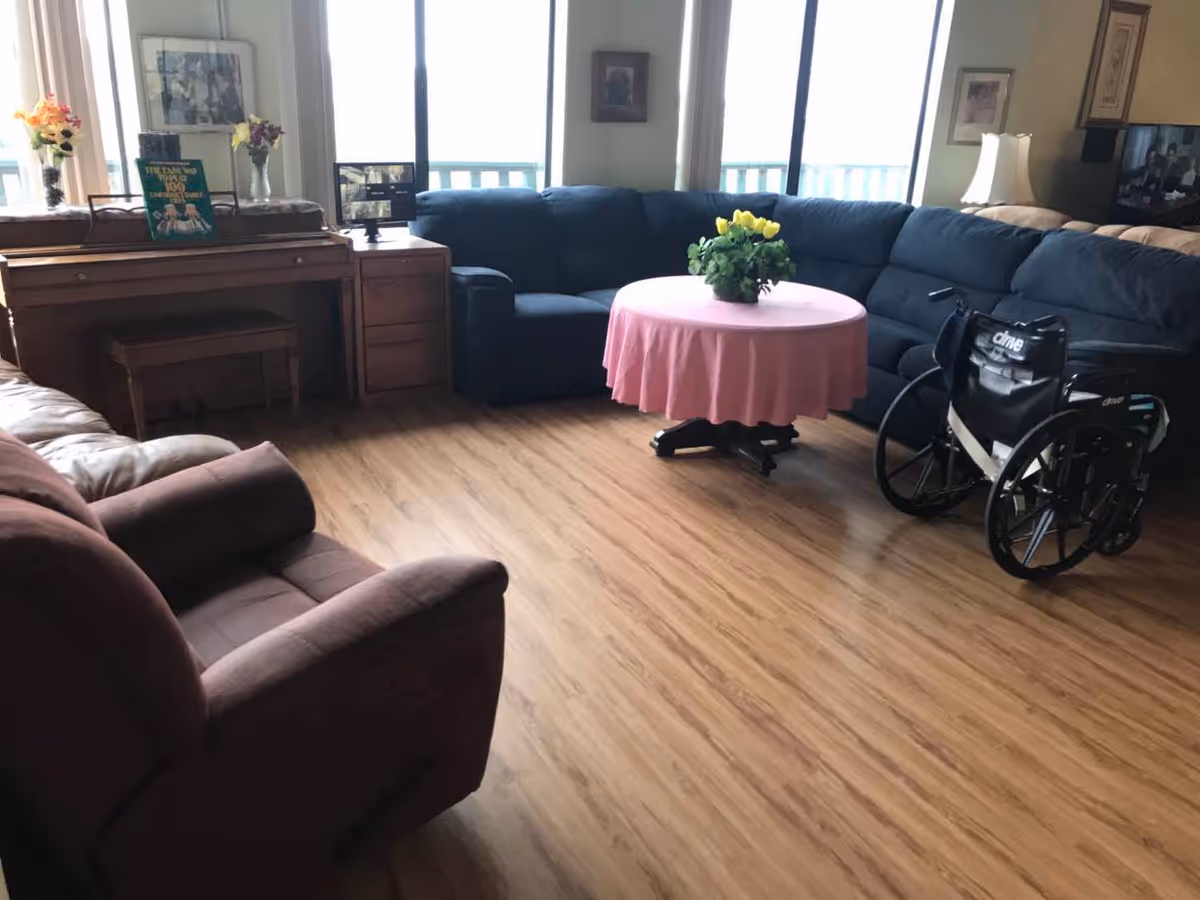 Bright senior living common area with blue sectional, pink round table, and wheelchair.