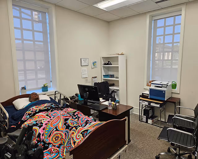 A patient bed with a person under a colorful patterned blanket in a small office-like room.