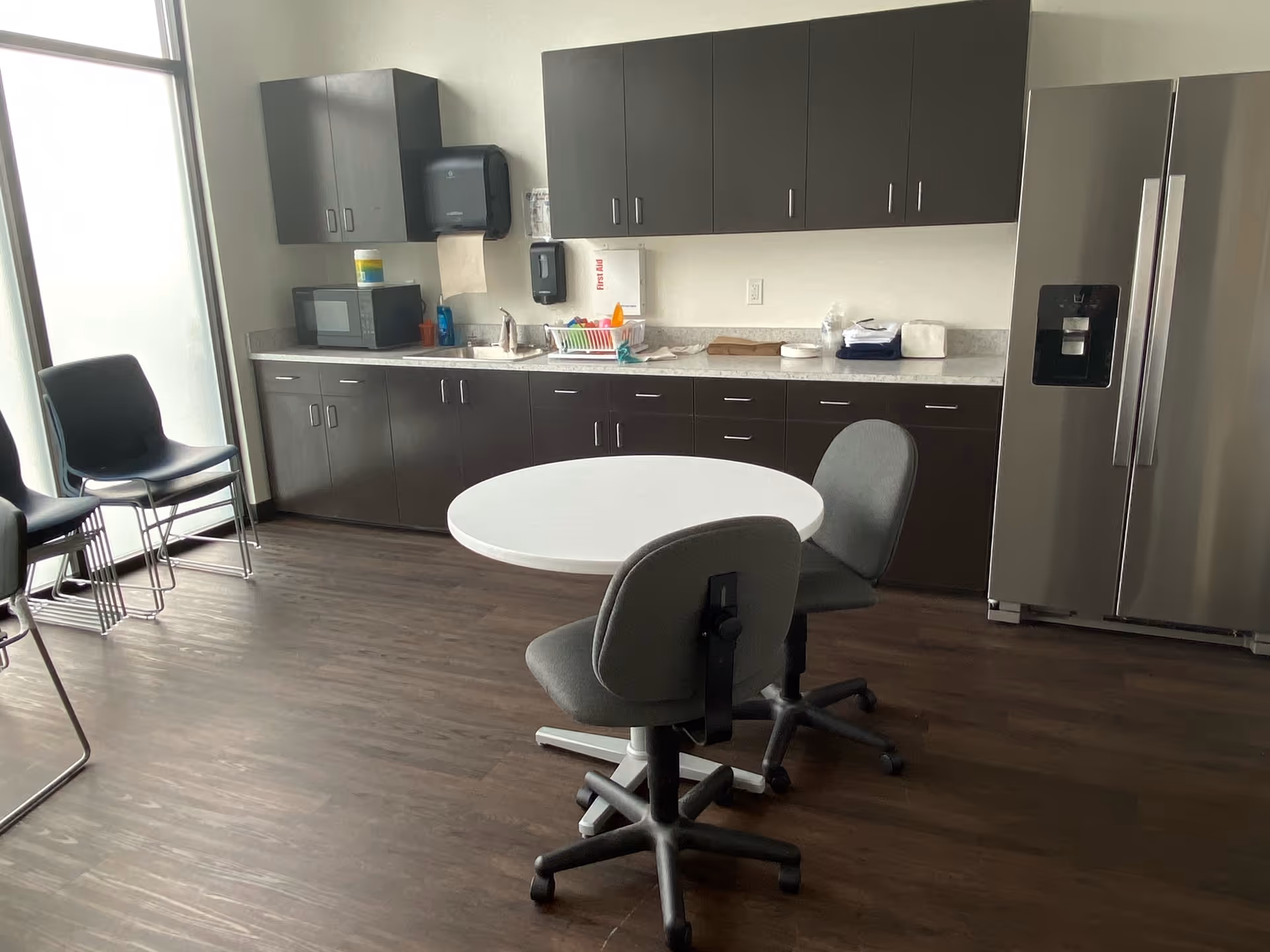 Modern kitchenette with dark cabinets, stainless fridge, round table, and chairs in a senior living facility.