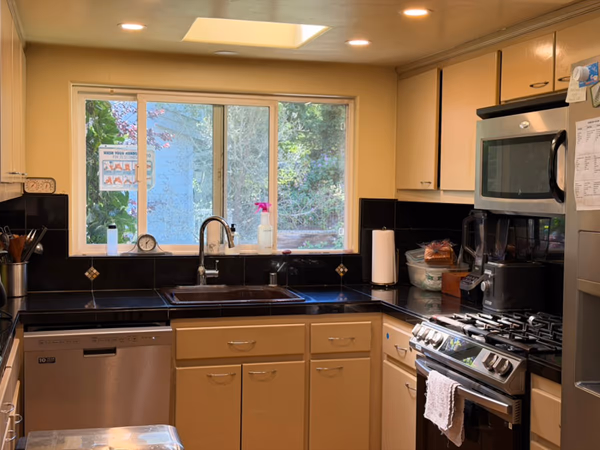 Kitchen with beige cabinets, black countertops, stainless steel stove, sink, microwave, and window view.