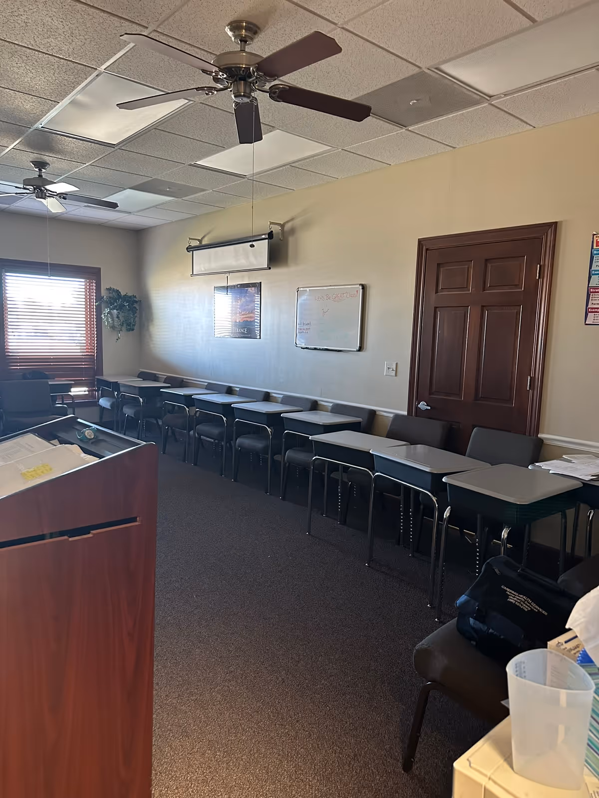 A classroom-style interior with a podium, row of desks, whiteboards, and ceiling fans.