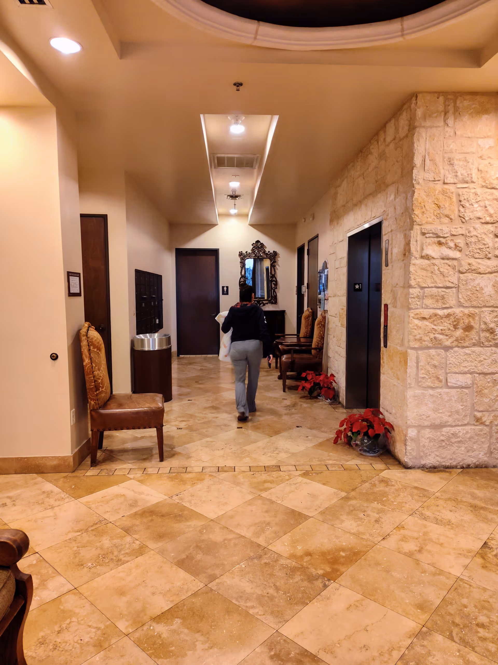 Interior hospital hallway with seating, elevator doors, stone wall, and a person walking away.