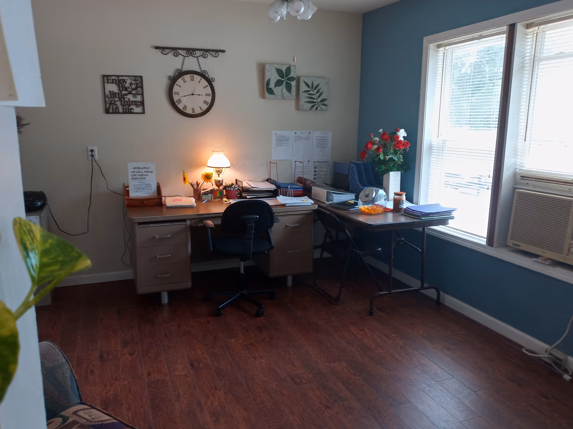 Office area with desks, a clock, lamp, paperwork, plants, and a window air conditioner.