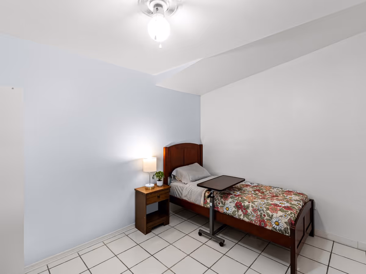 Small furnished bedroom with single bed, nightstand, lamp, plant, and tiled floor.