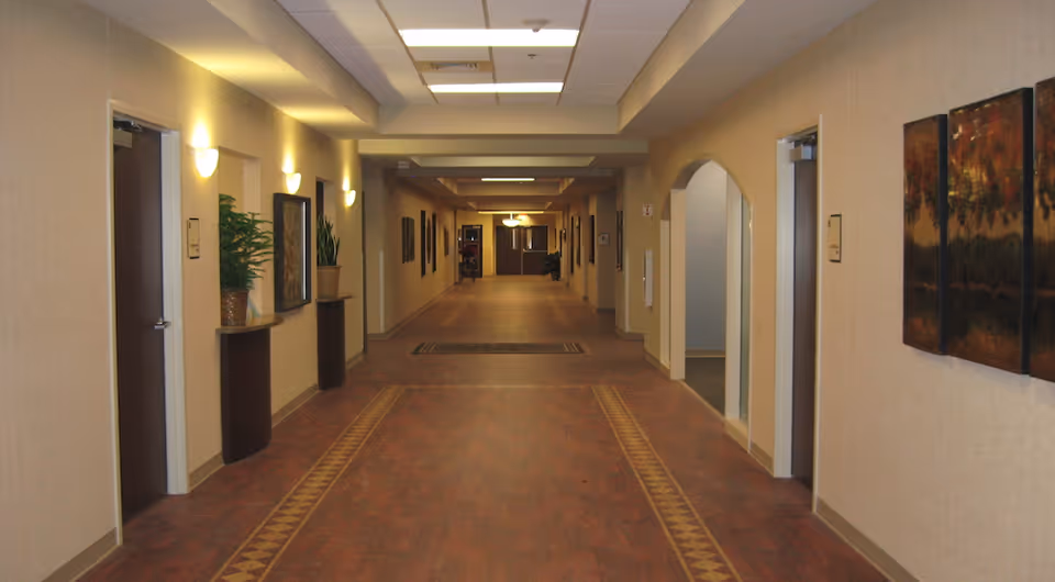 Long indoor corridor in a care facility with doors, art, and potted plants.