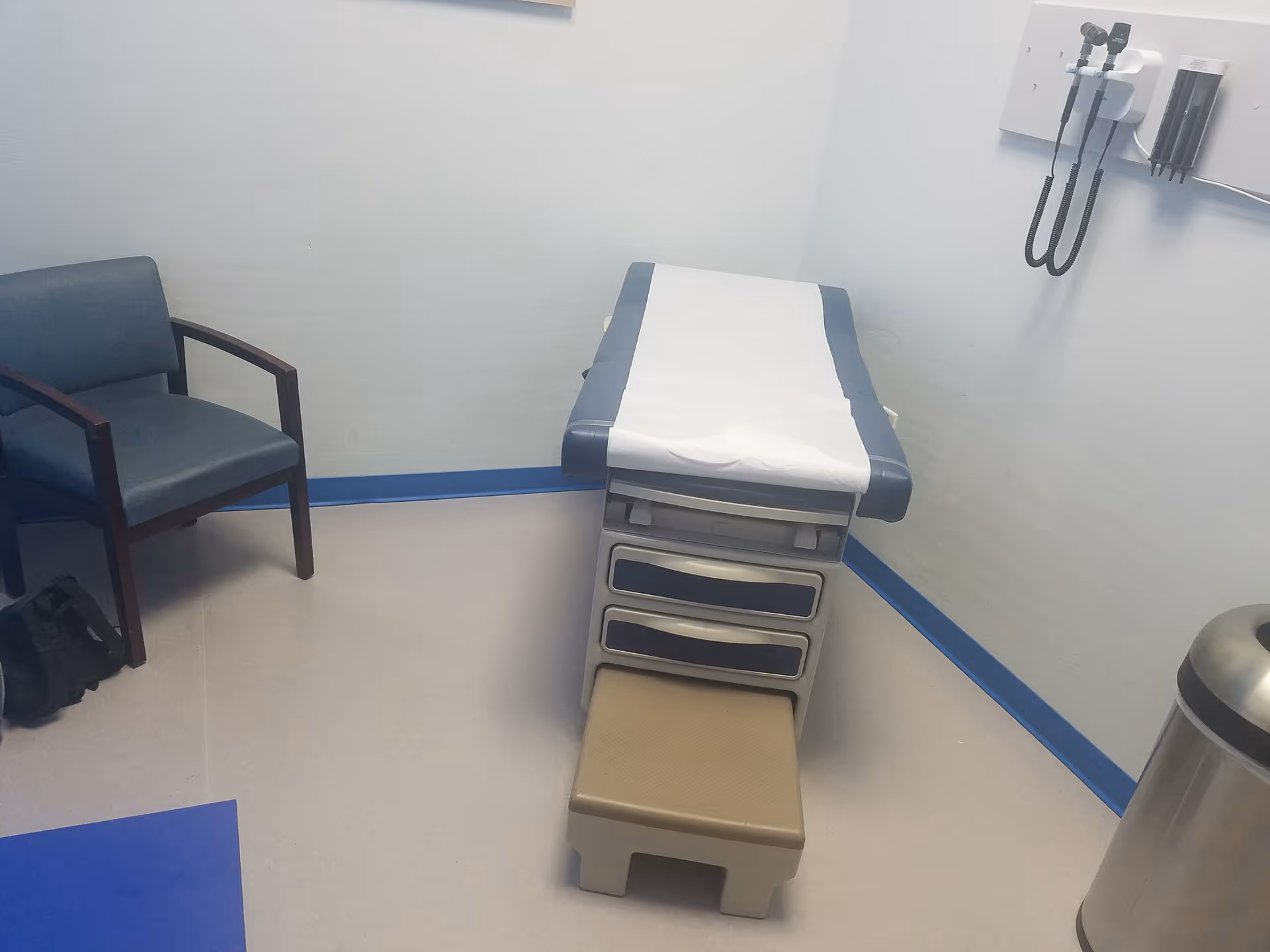 Medical examination room with a padded exam table, chair, and wall-mounted medical equipment.