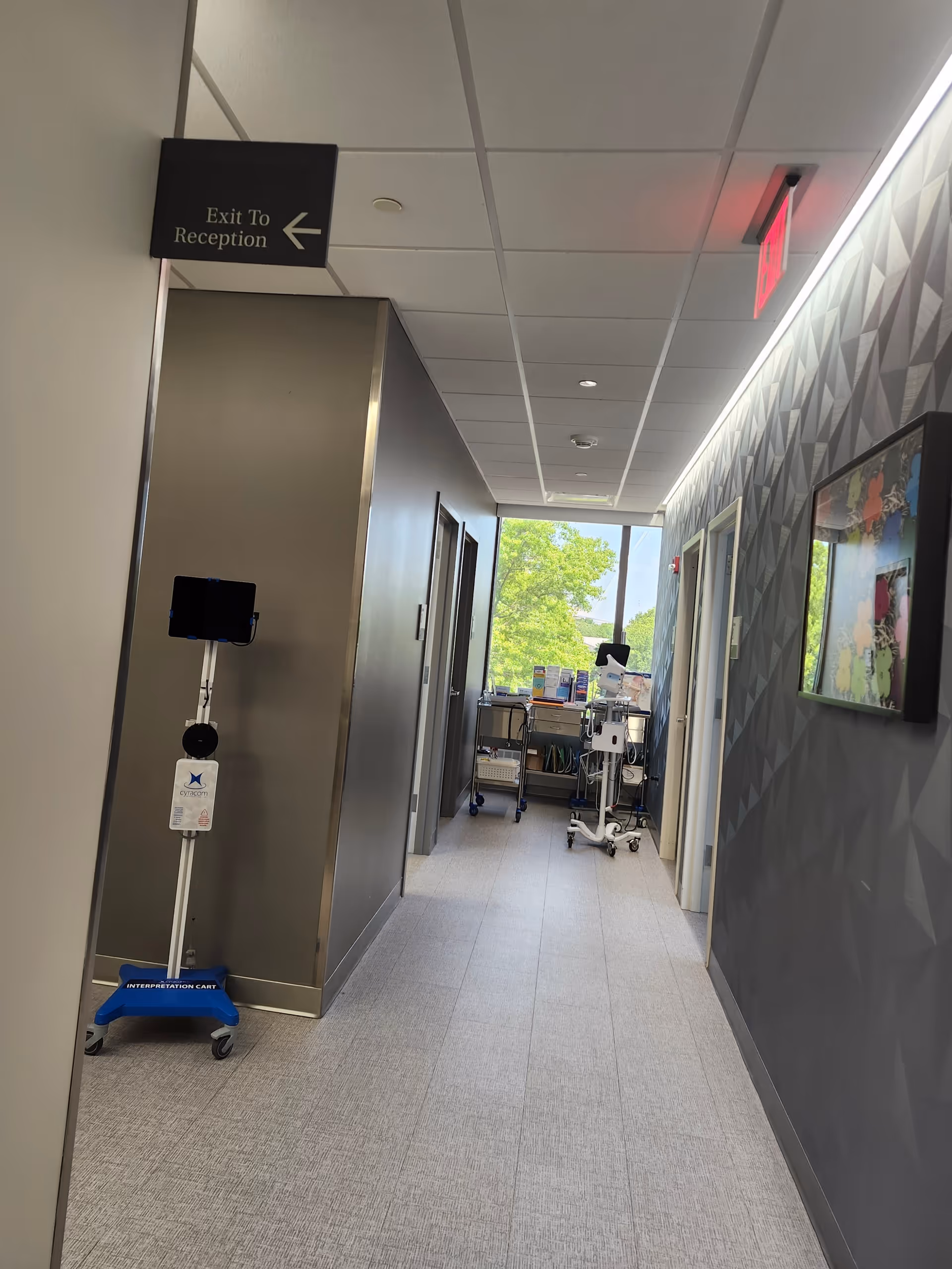 Hallway inside a facility with an exit sign, medical equipment cart, and patterned wall.