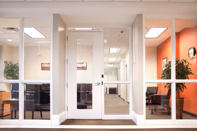 Glass-walled hallway with a central door, seating, plants, and orange accent wall.