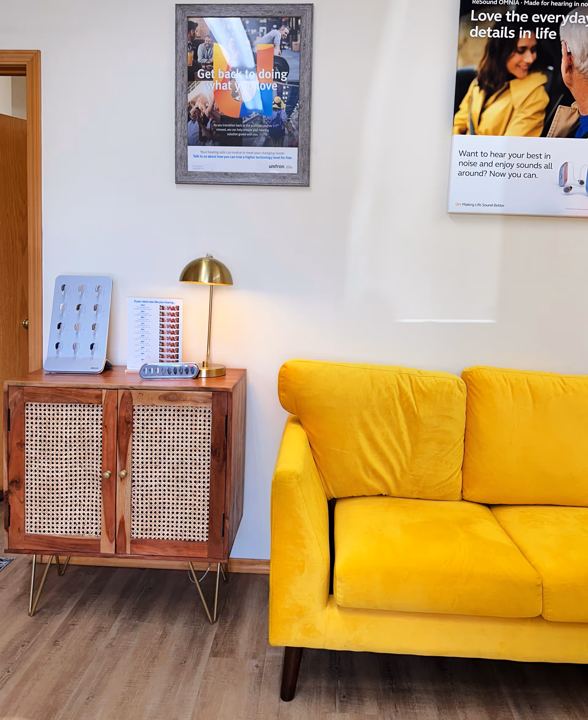 Bright waiting area with a yellow sofa, wooden cabinet, lamp, and framed posters.