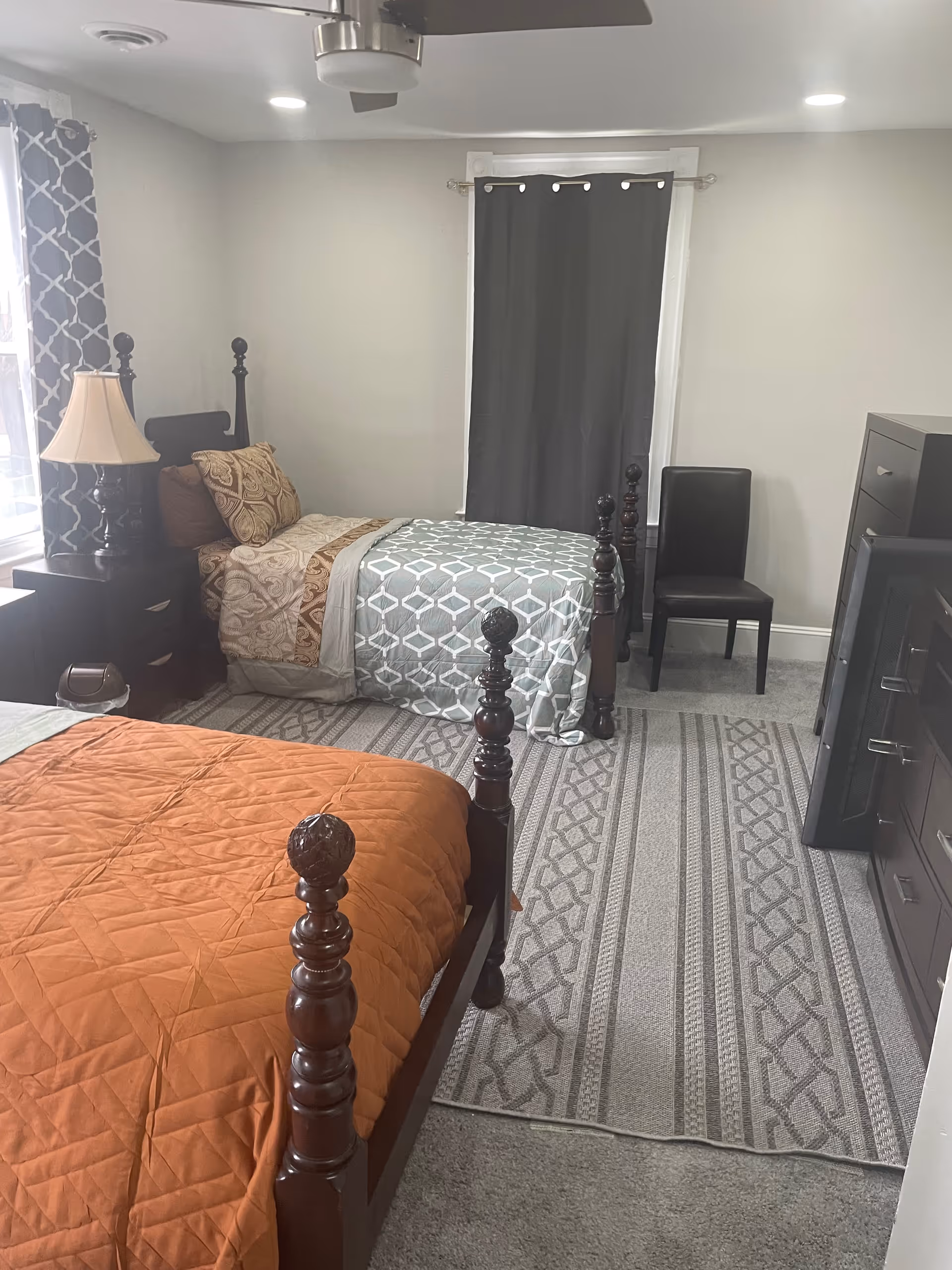 Two wooden four-poster beds in a bright bedroom with a nightstand, lamp, dresser, and curtains.