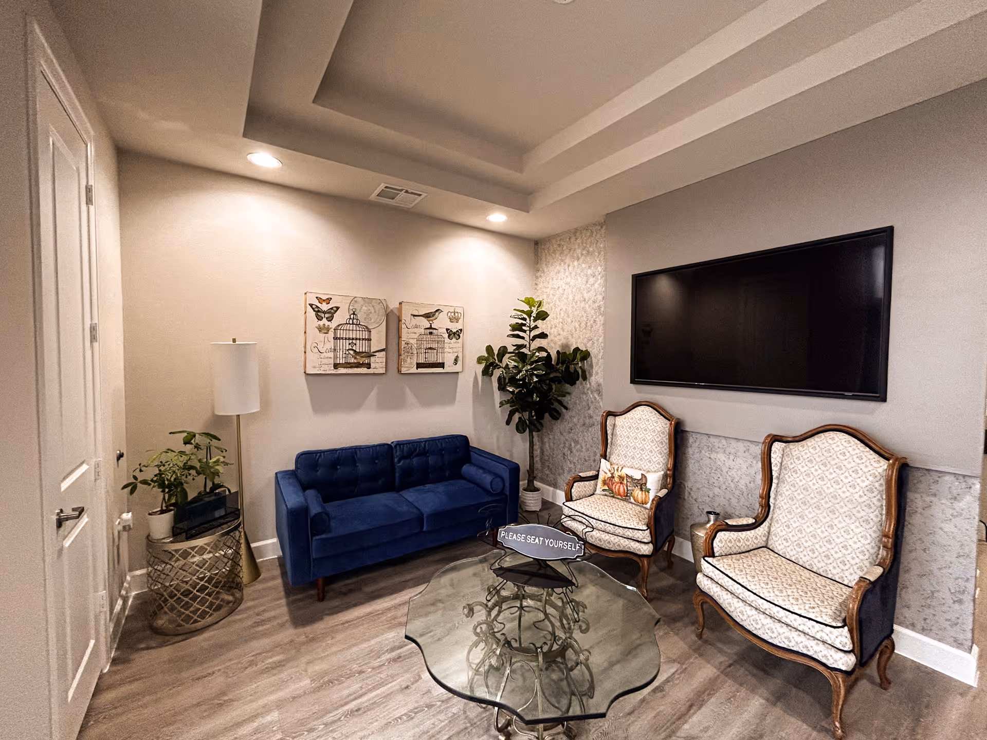 Modern lounge area with blue sofa, two ornate chairs, glass coffee table and wall-mounted TV.