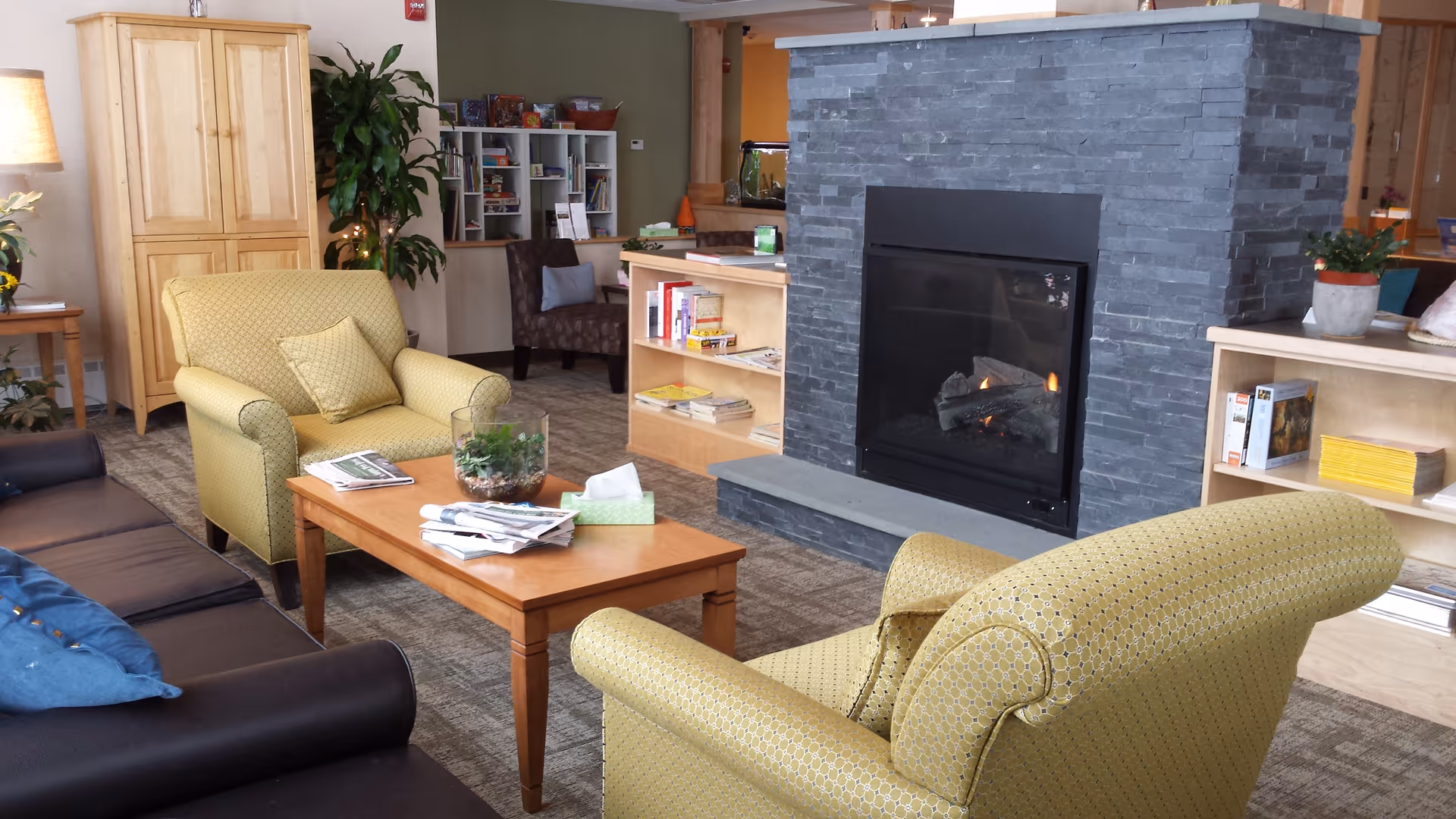 Cozy facility living room featuring a stone fireplace, yellow upholstered chairs, and a wooden coffee table.