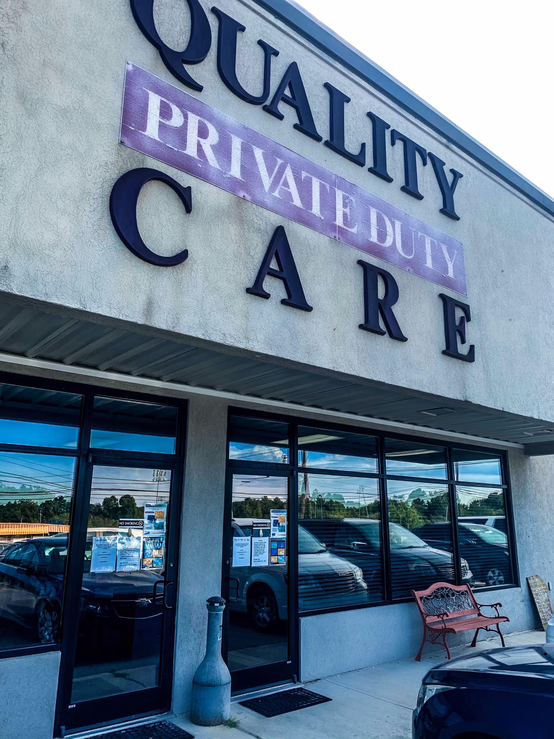 Exterior view of Quality Private Duty Care storefront with glass doors and a bench.