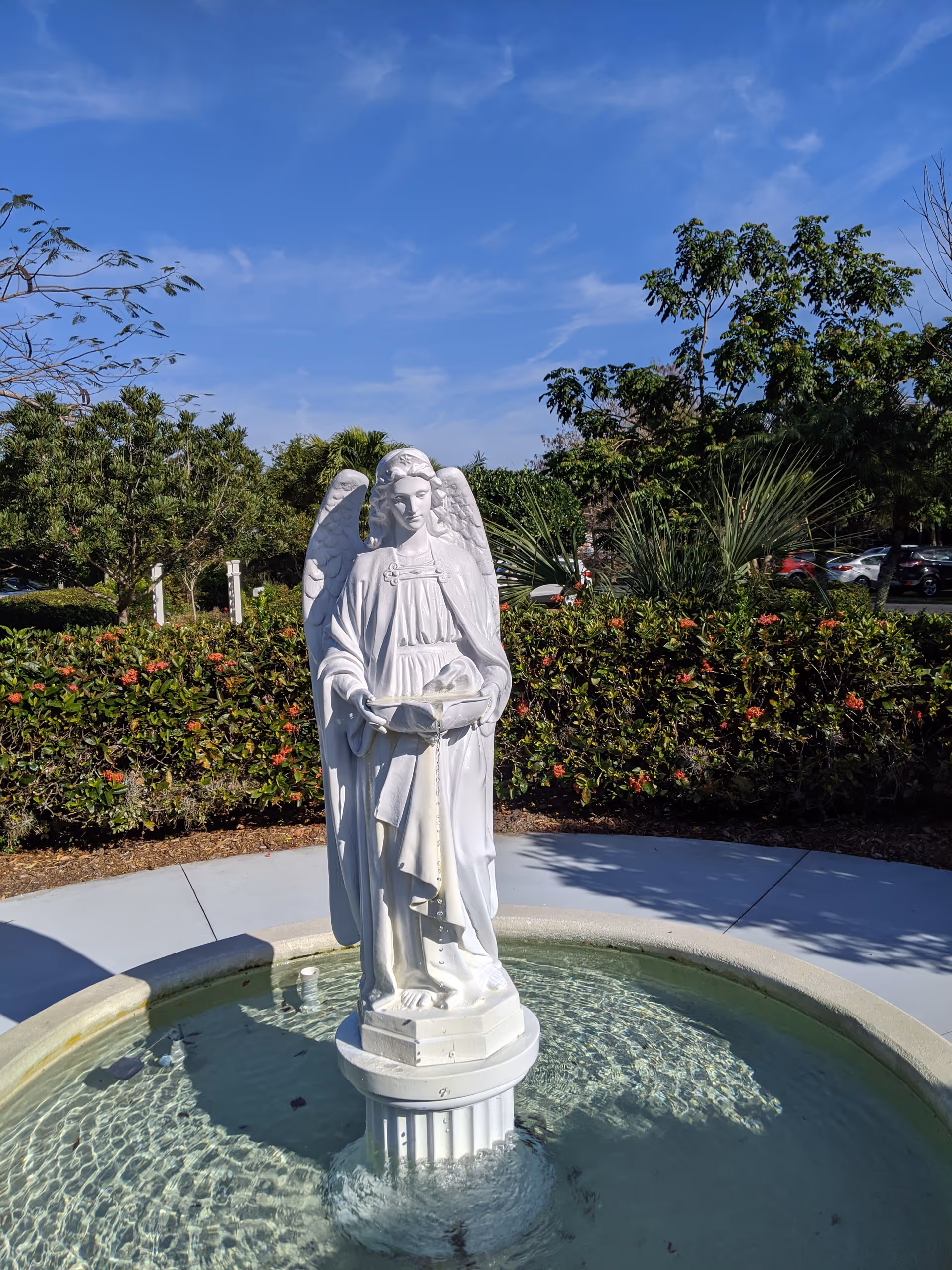 White angel statue stands in a circular fountain with greenery and blue sky behind.