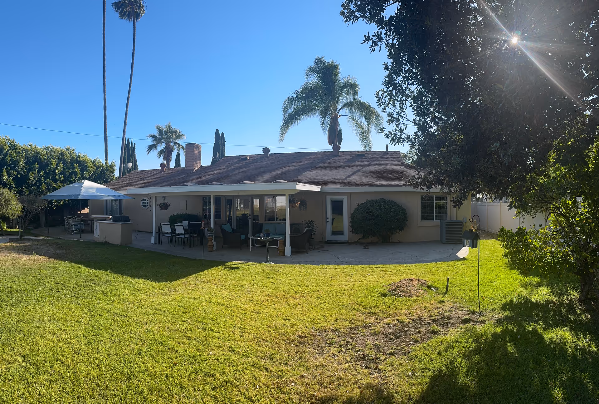 Single-story house with a covered patio, outdoor seating, and palm trees in a sunny yard.