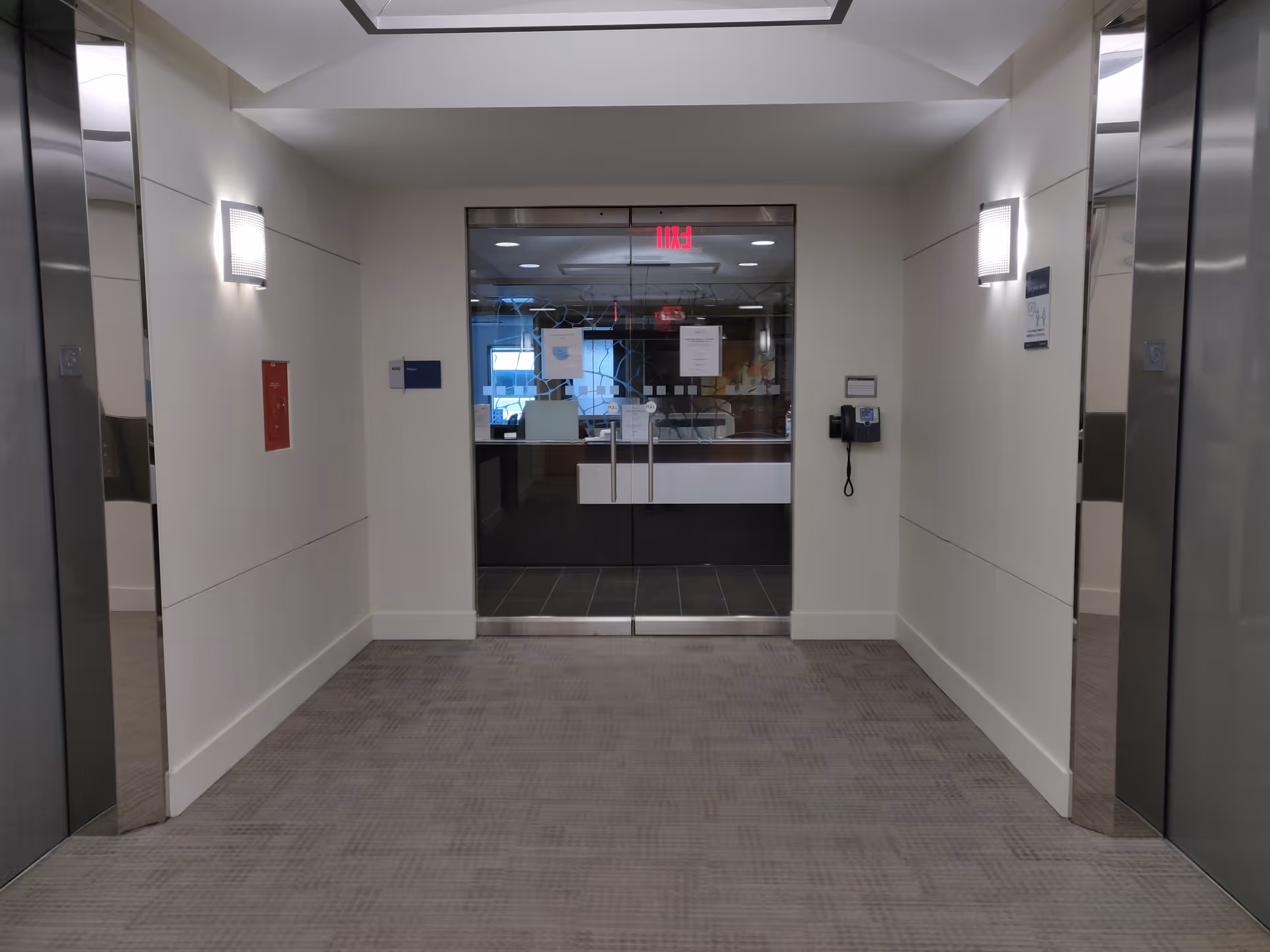 Bright hallway with glass entry, adjacent elevators, and a reception desk beyond.
