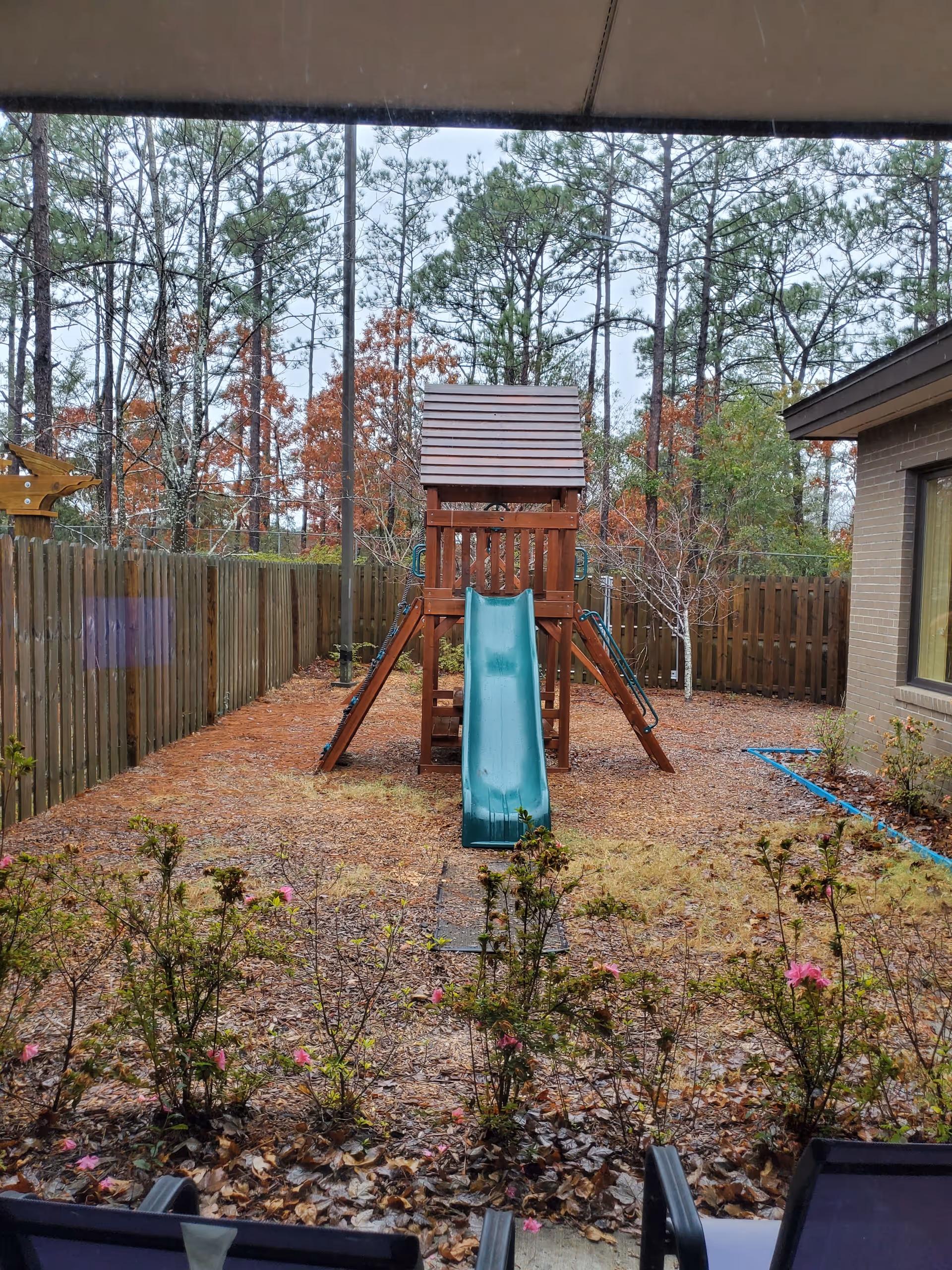 Wooden playset with teal slide in a fenced backyard near a brick house