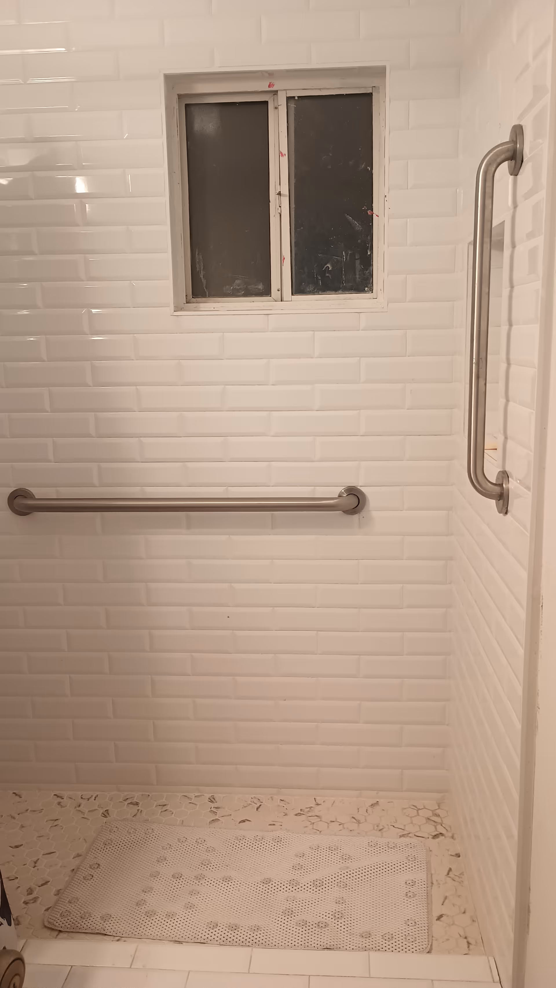 White tiled bathroom with grab bars, small window, and textured mat.