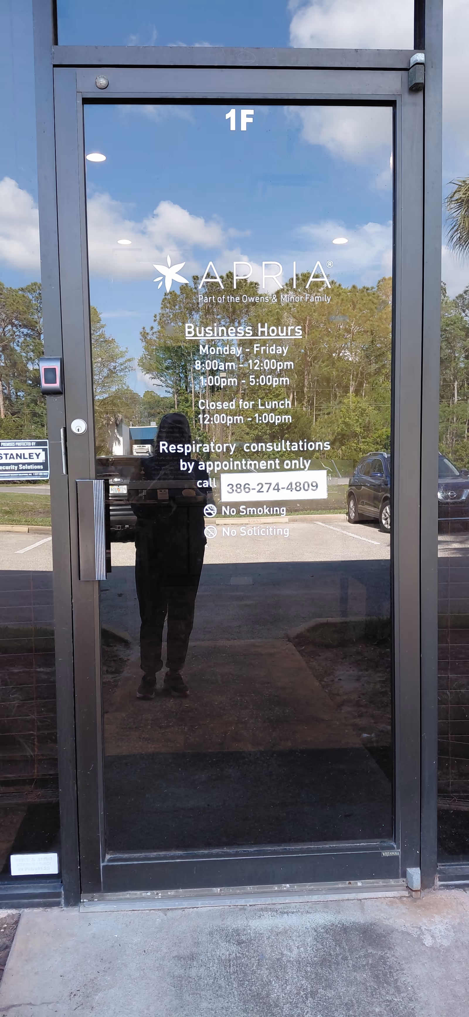 Glass door entrance with Apria Healthcare logo, business hours, and reflections of trees.