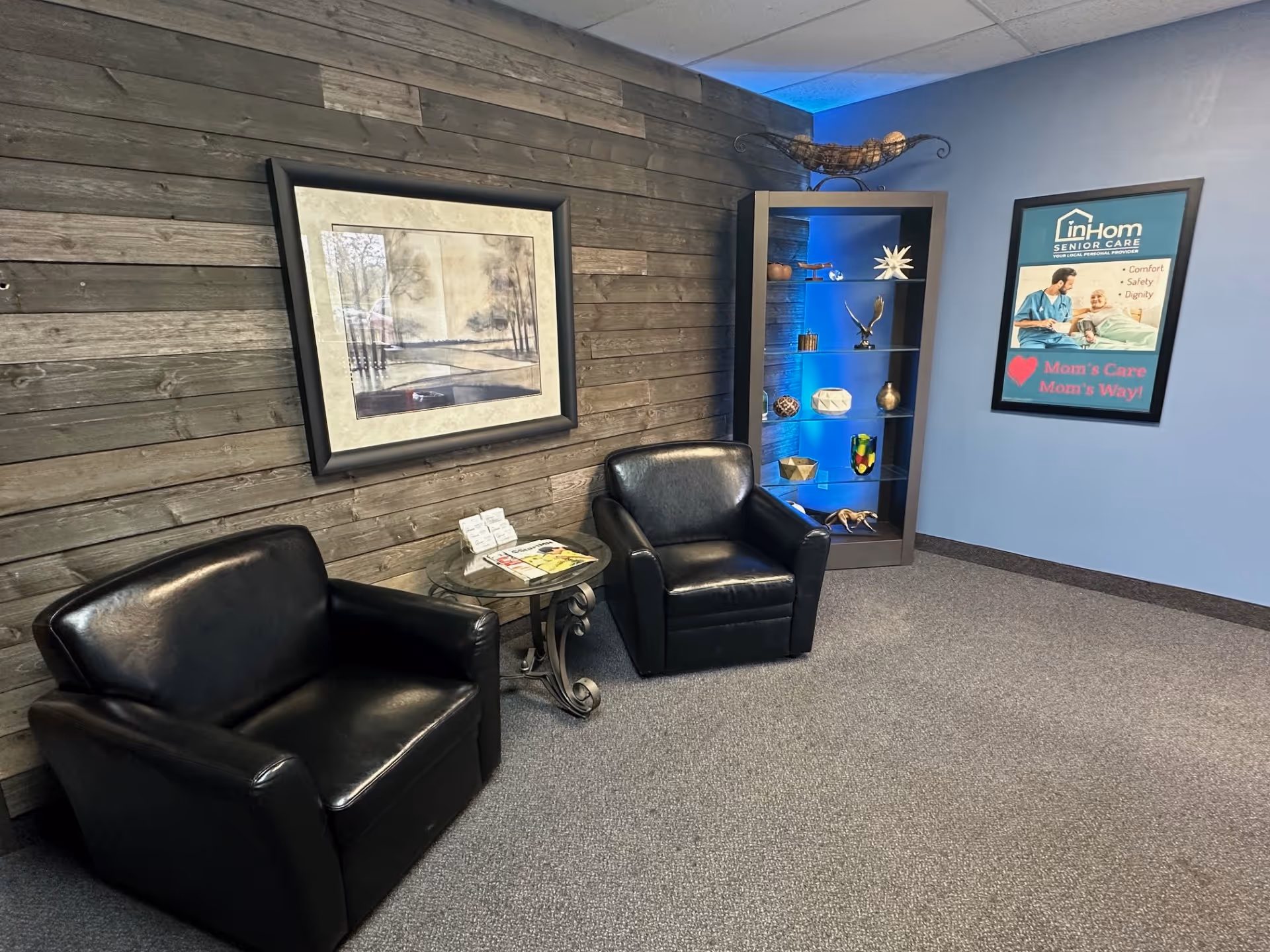 Lounge area with two black leather chairs, glass table, wood wall, and blue display cabinet.