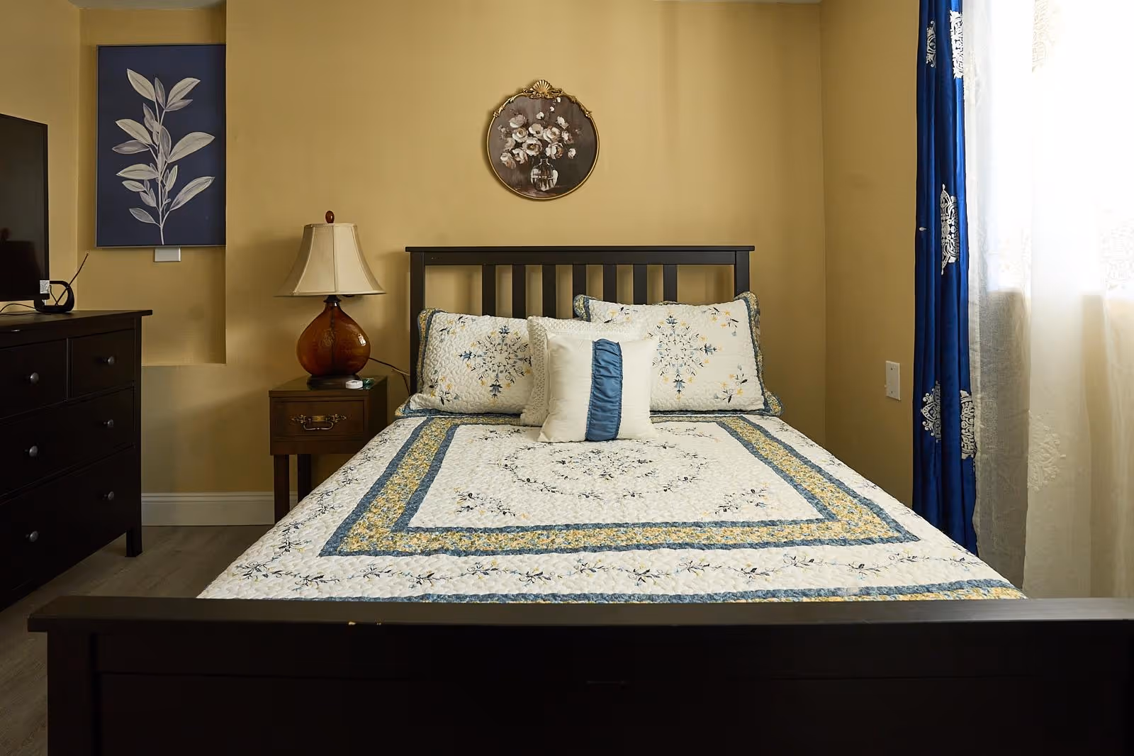 A neatly made bedroom with a dark wood bed, nightstand, lamp, dresser, and blue curtains.
