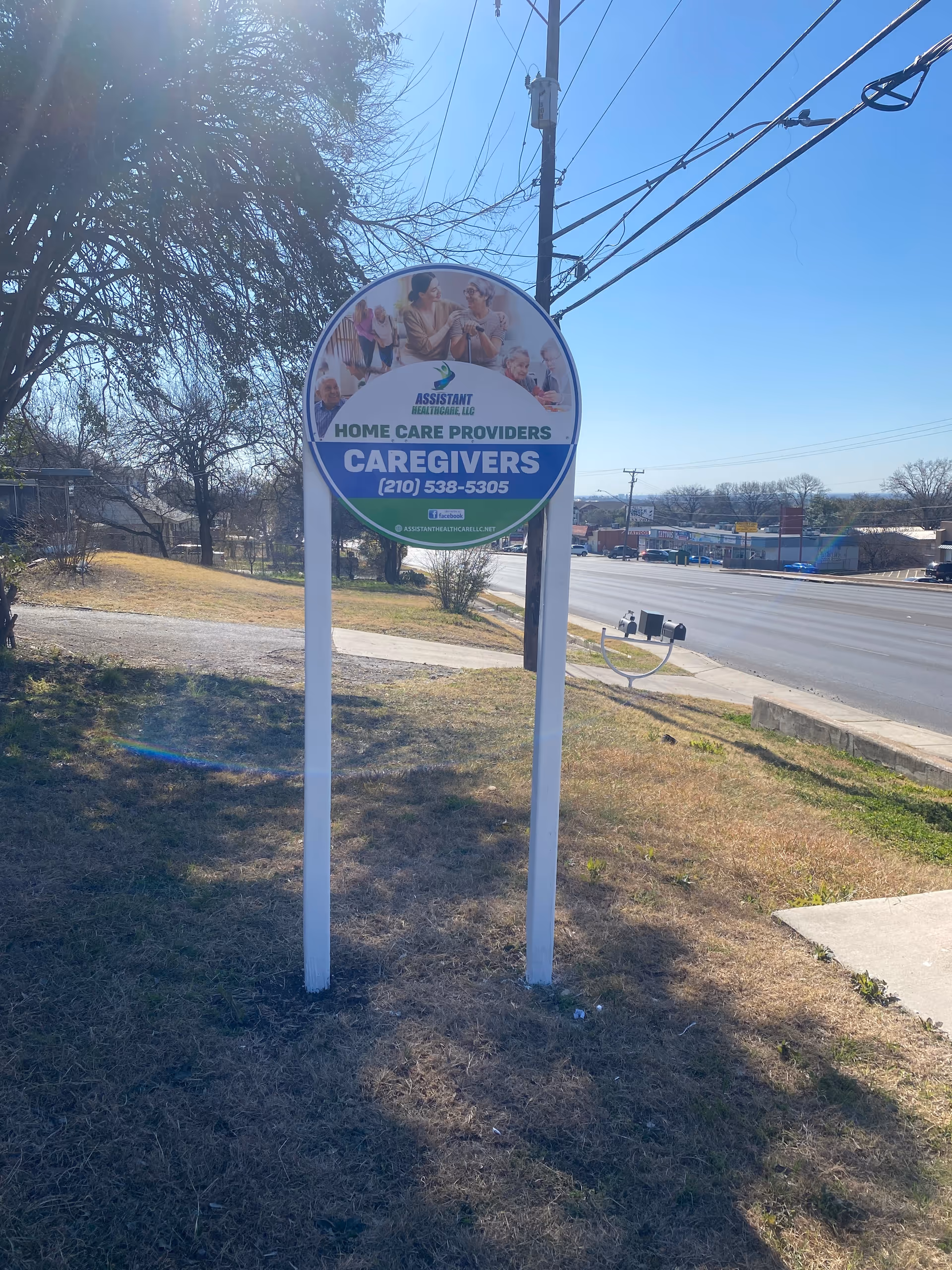 outdoor circular sign on two posts advertising caregiver services from Assistant Healthcare LLC beside a street