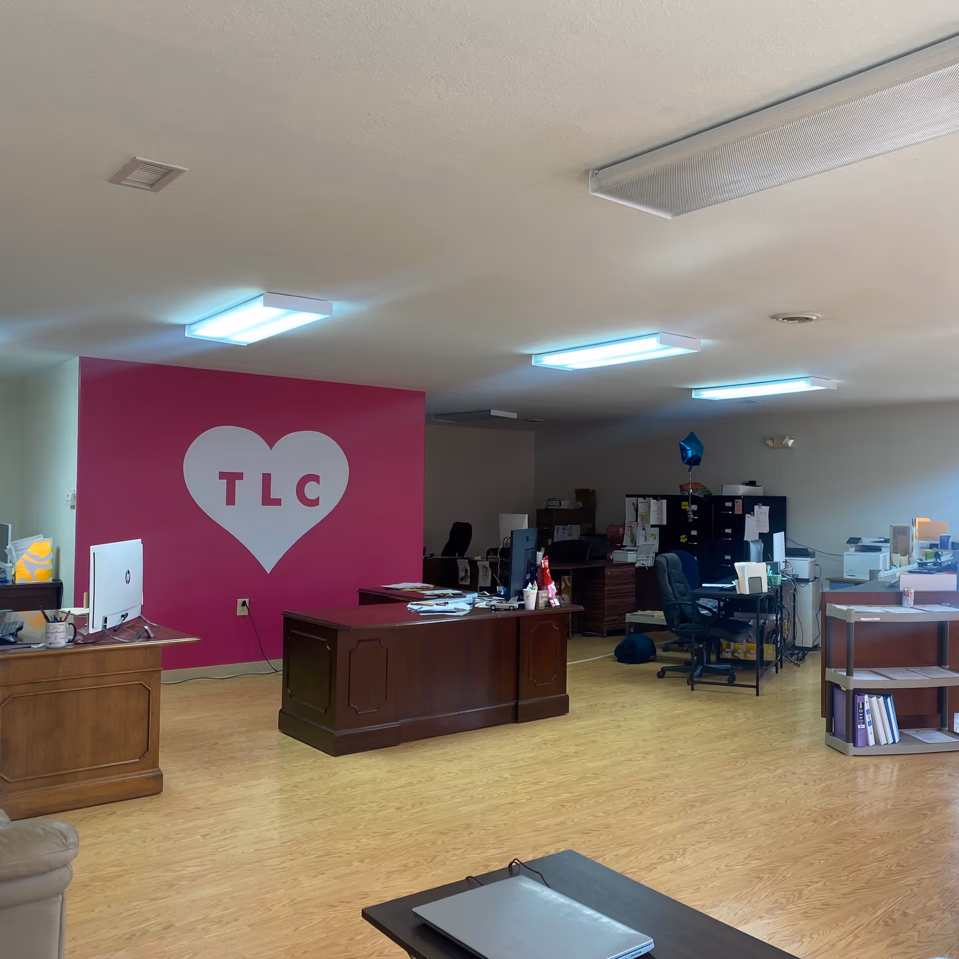 Office reception area with pink TLC heart wall, desks, computers, and filing cabinets.