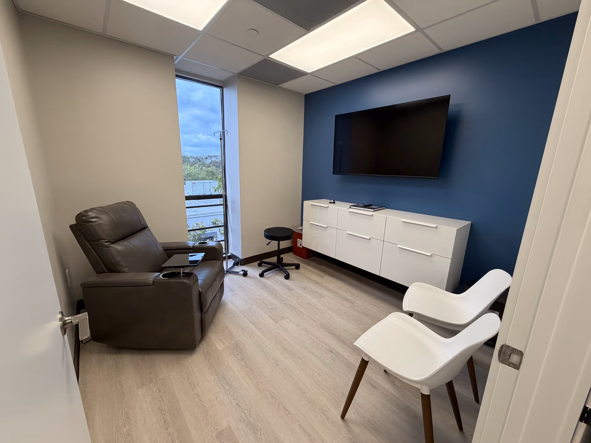 Small modern living room with blue accent wall, TV, cabinet, leather chair, and white chairs.