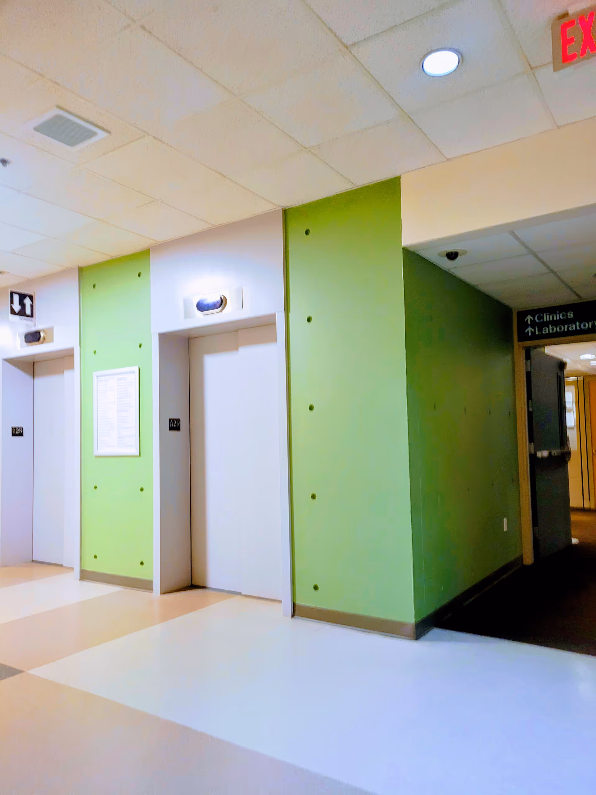 Interior hospital corridor with green walls, elevators, and directional signs along a tiled floor.