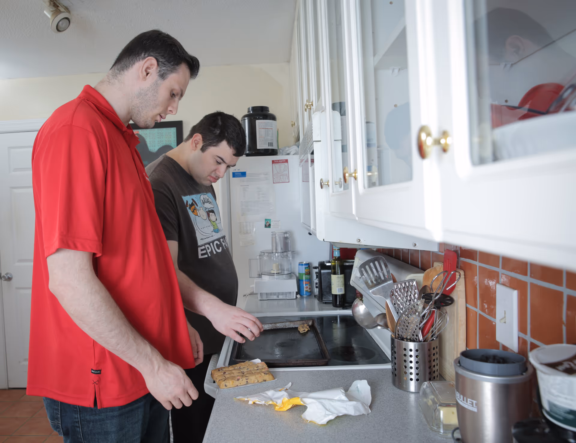 Two men stand by a stove in a compact kitchen with utensils.