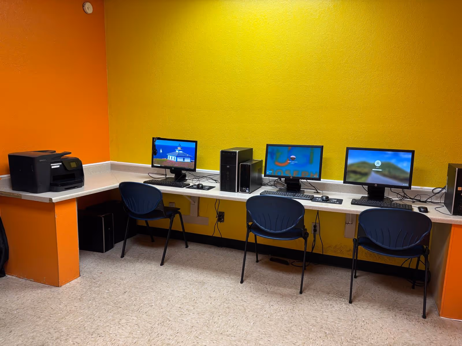 Interior computer lab with four desktop stations, printers, and chairs along a bright yellow wall.