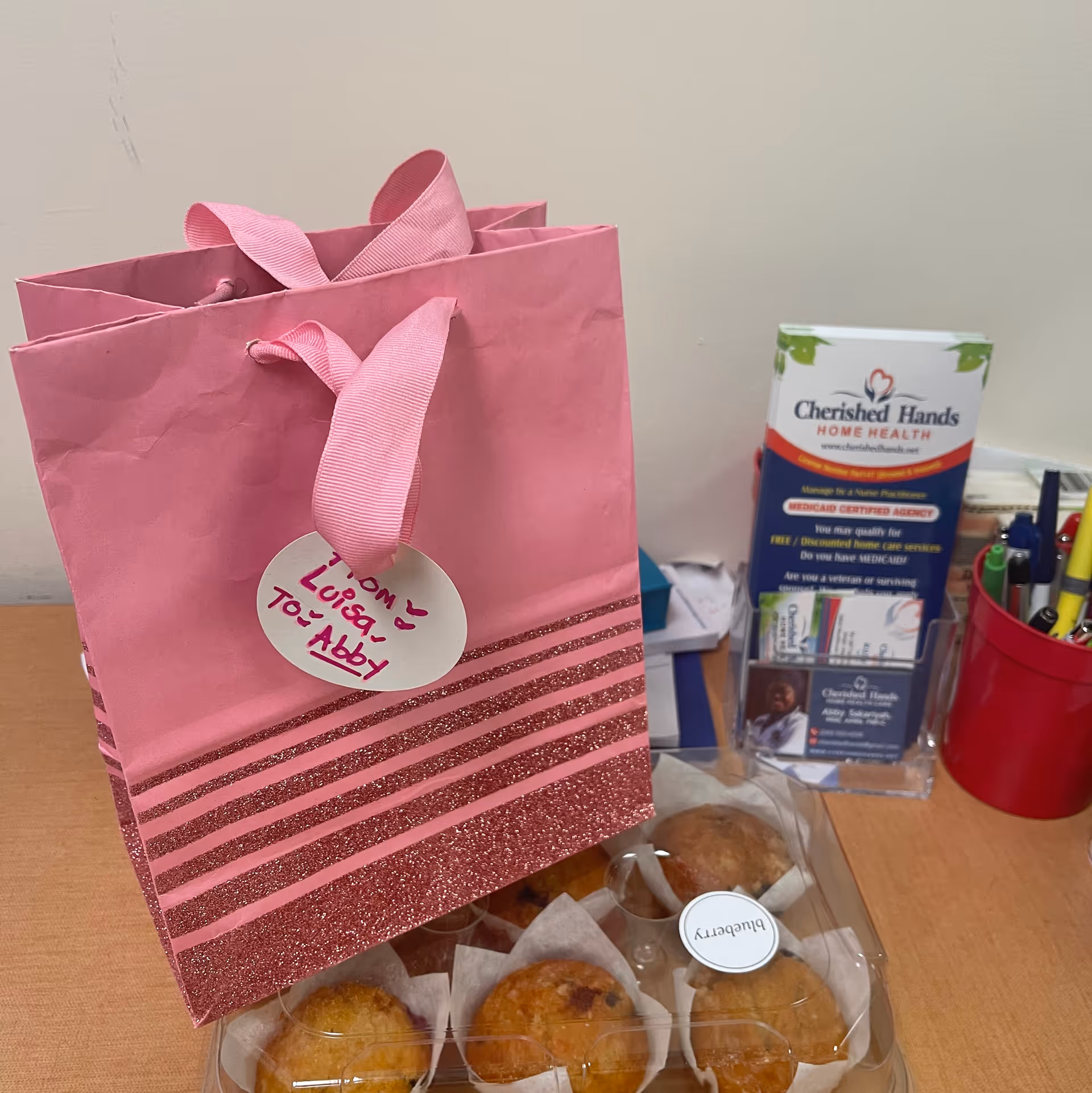 Pink gift bag with muffins and Cherished Hands Home Health brochure on a desk.