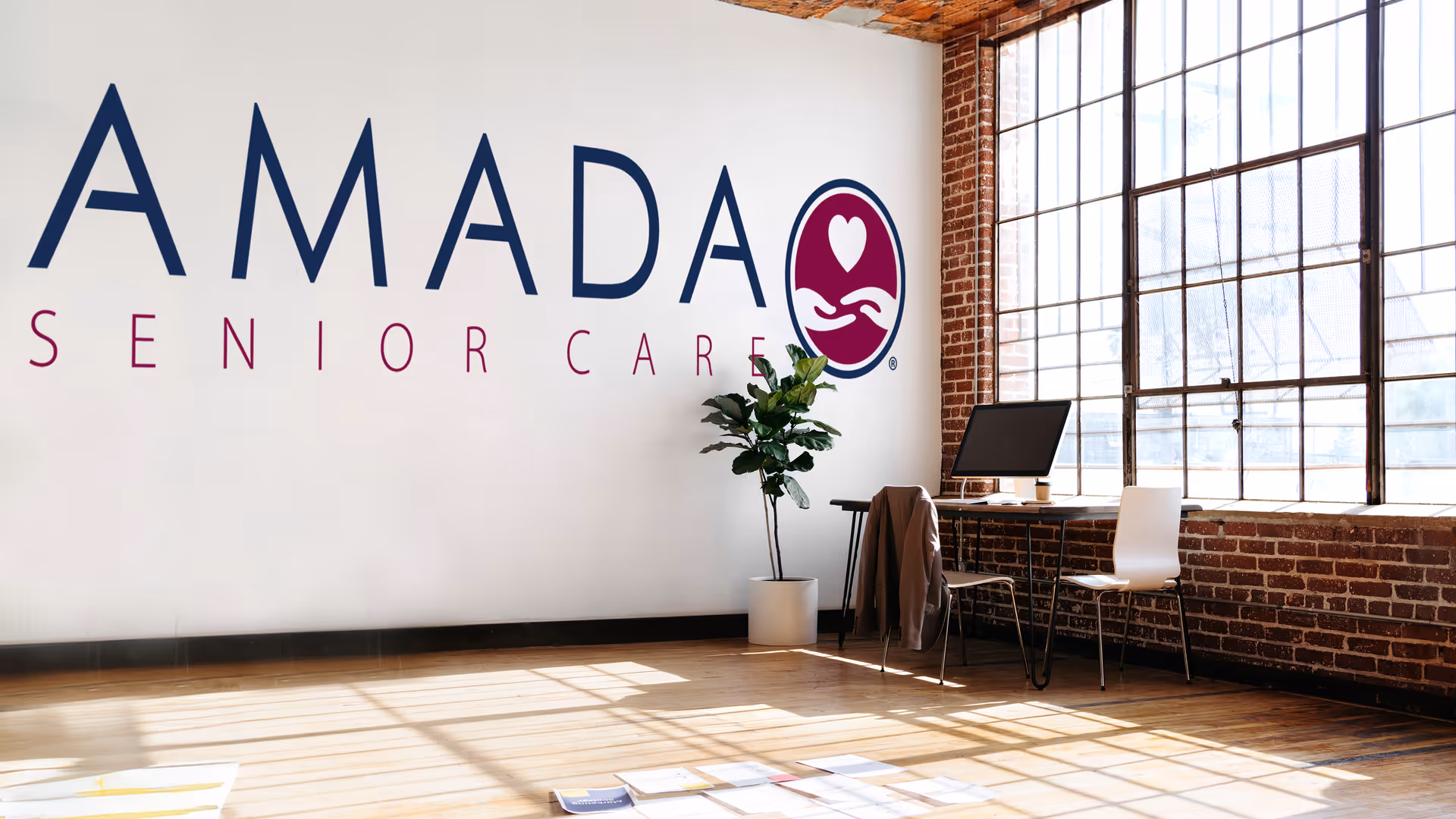 Amada Senior Care lobby area with logo, desk, chair, plant, and brick-framed windows.