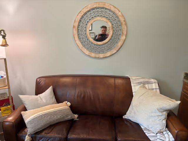 Living room scene with a brown leather sofa, pillows, and a round wall mirror.