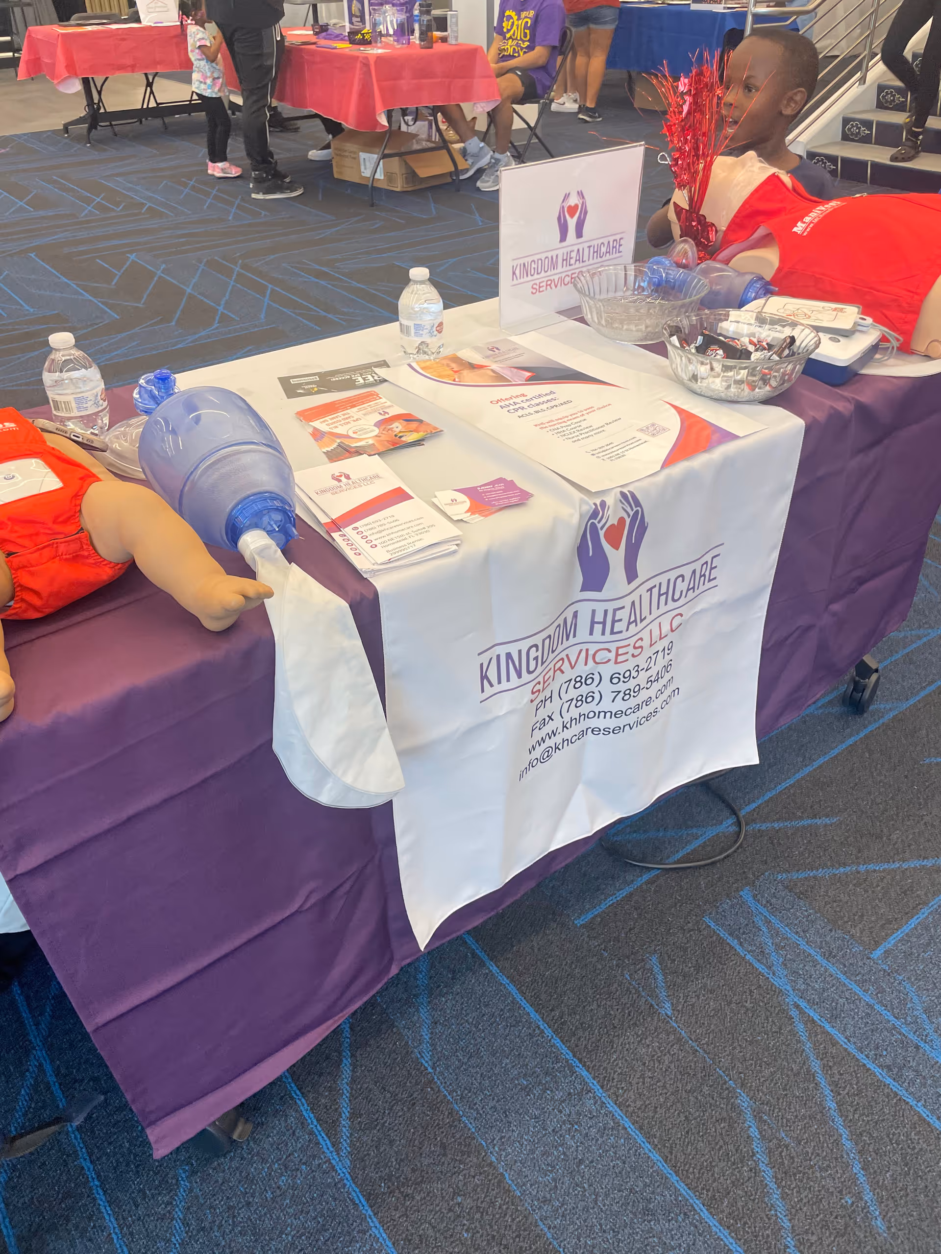 Indoor health fair booth with Kingdom Healthcare Services banner and brochures on a purple tablecloth.