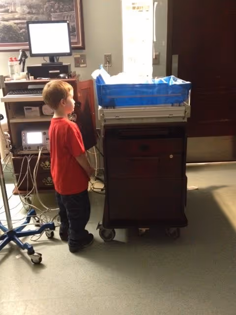 A young child stands beside a medical cart and monitors in a hospital room.