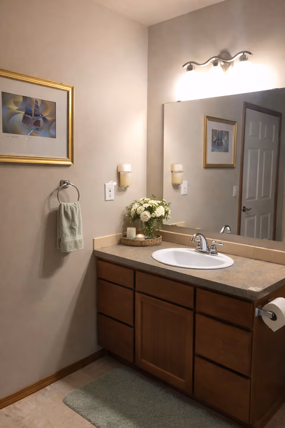 A bathroom vanity with sink, mirror, flowers, and soft beige walls in a senior home.